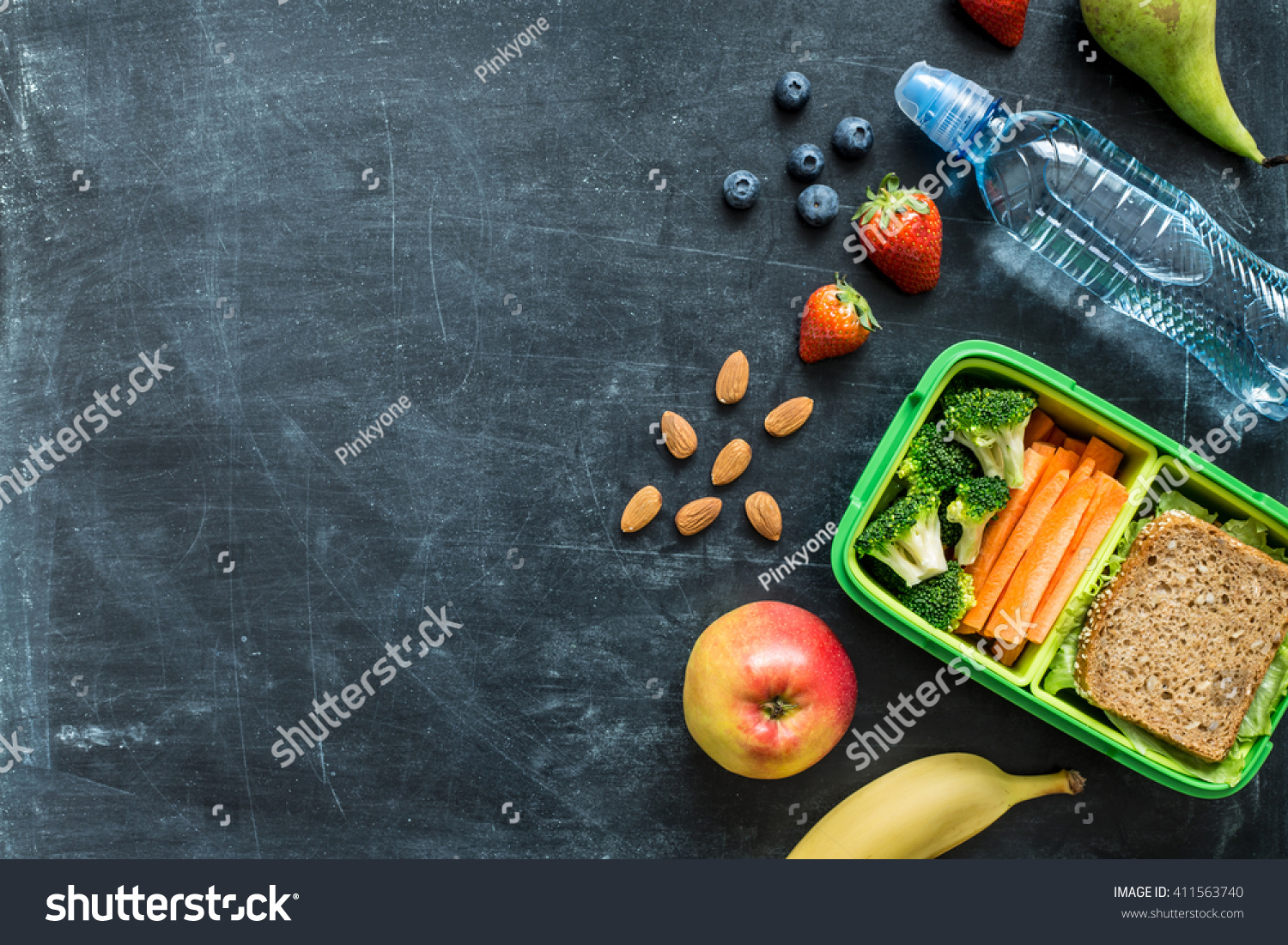 School lunch box with sandwich, vegetables, water, almonds and fruits on black chalkboard. Healthy eating habits concept - background layout with free text space. Flat lay composition (top view). #411563740