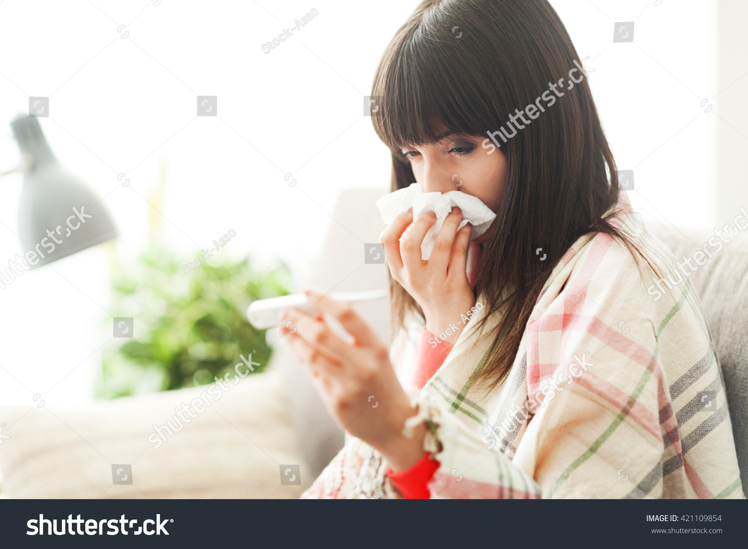 Young sick woman with cold and flu, she is blowing her nose and measuring her body temperature #421109854