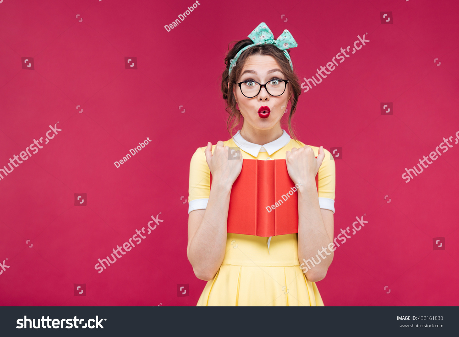 Charming surprised pinup girl in glasses standing and holding red book over pink background #432161830