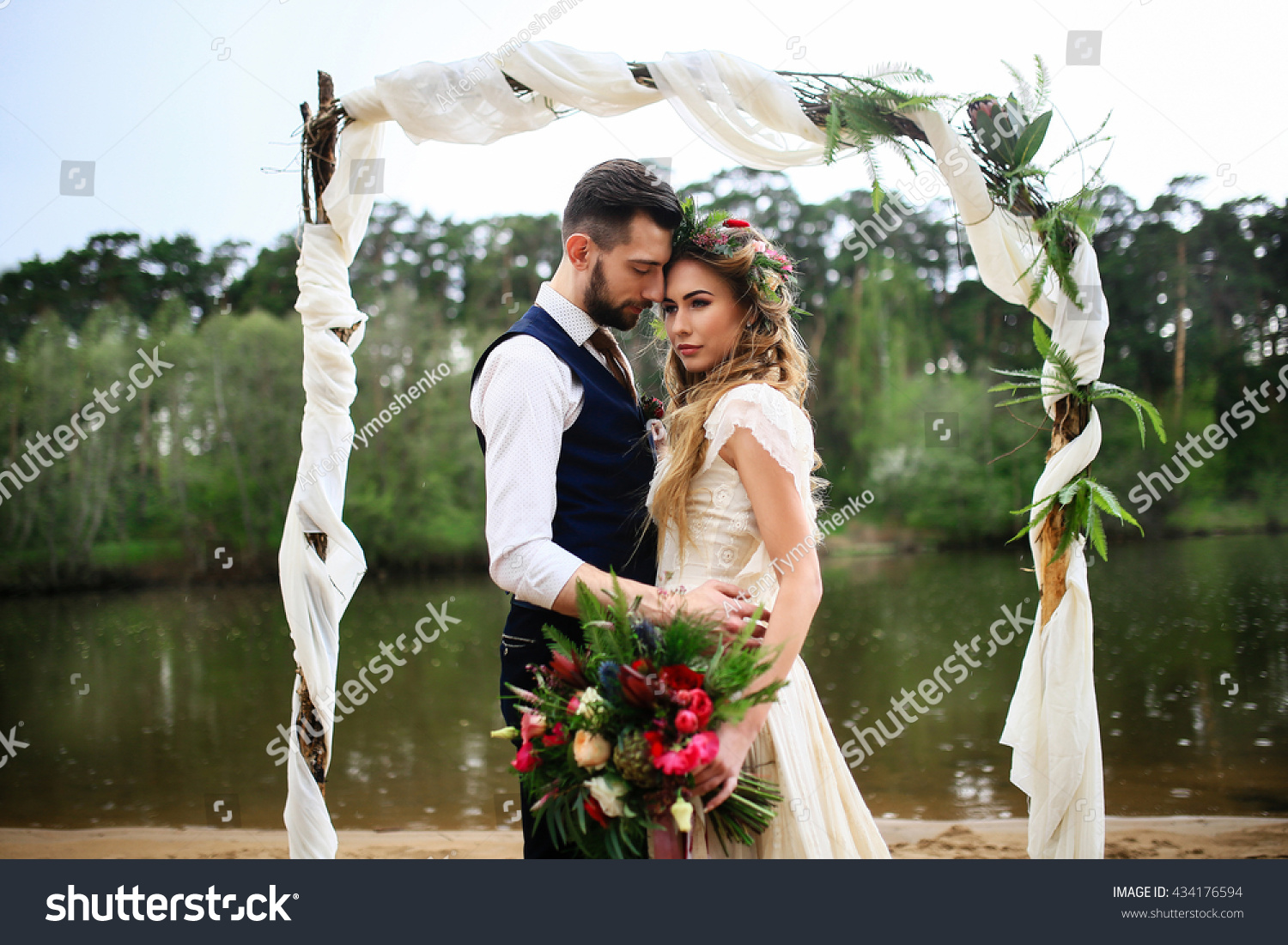 the groom in a wedding suit and boutonniere and bride in lace dress with bouquet and wreath , wedding arch on a background of lake #434176594
