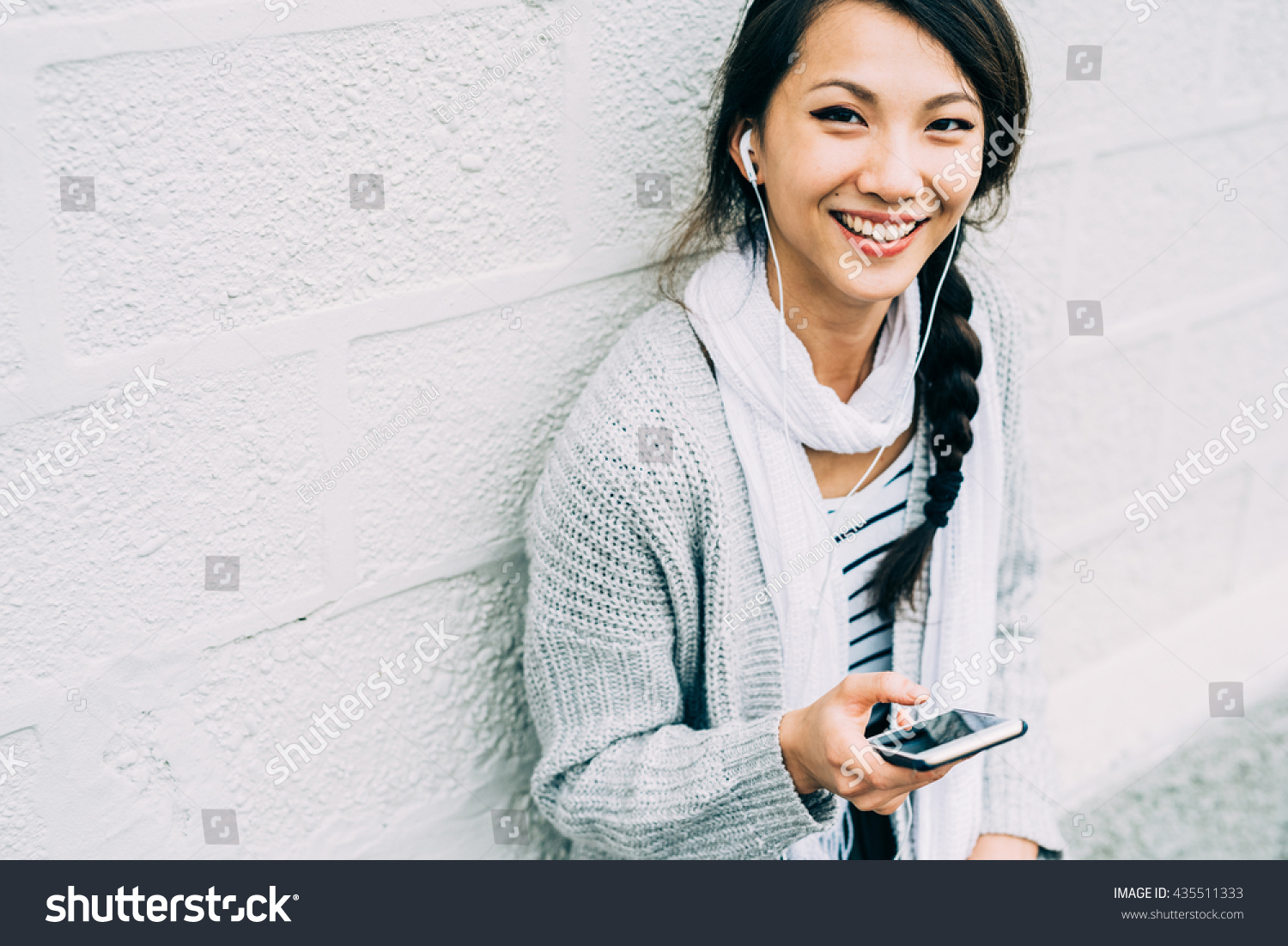 Half length of young beautiful asiatic woman holding a smart phone listening music with earphones, looking in camera, smiling - technology, happiness, music concept #435511333