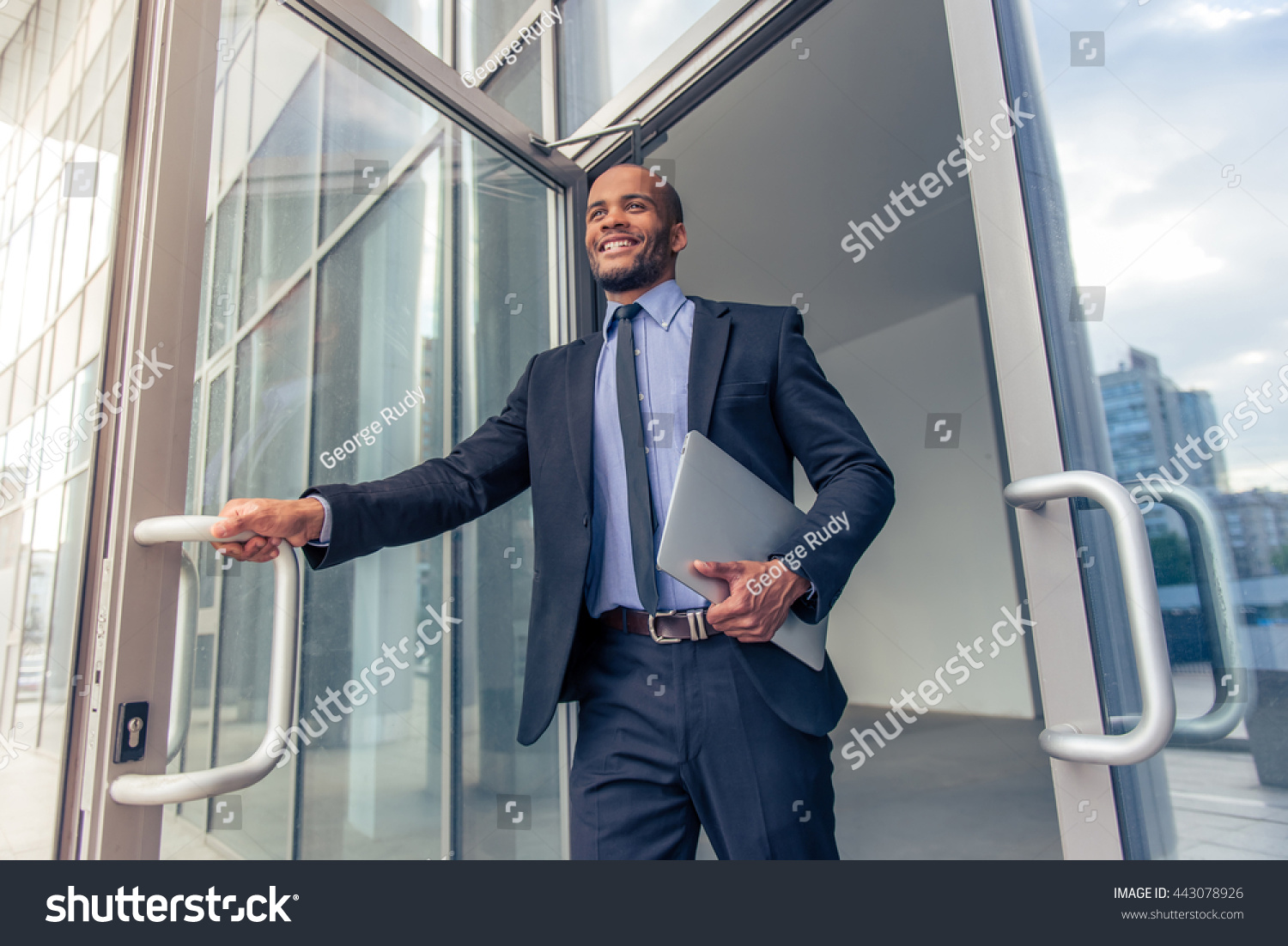 Low angle view of handsome young Afro American businessman in classic suit holding a laptop and smiling while leaving the office building #443078926