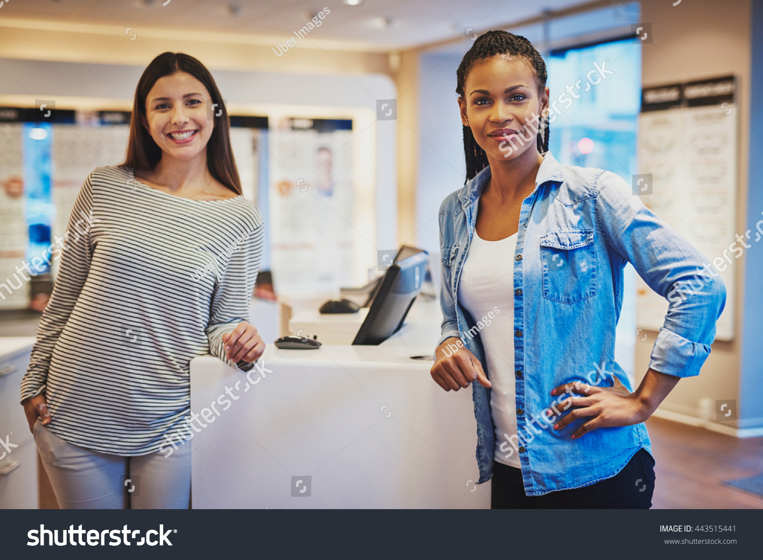 Smiling diverse girl friends wearing striped blouse and blue jean shirt stand against counter in optical shop #443515441