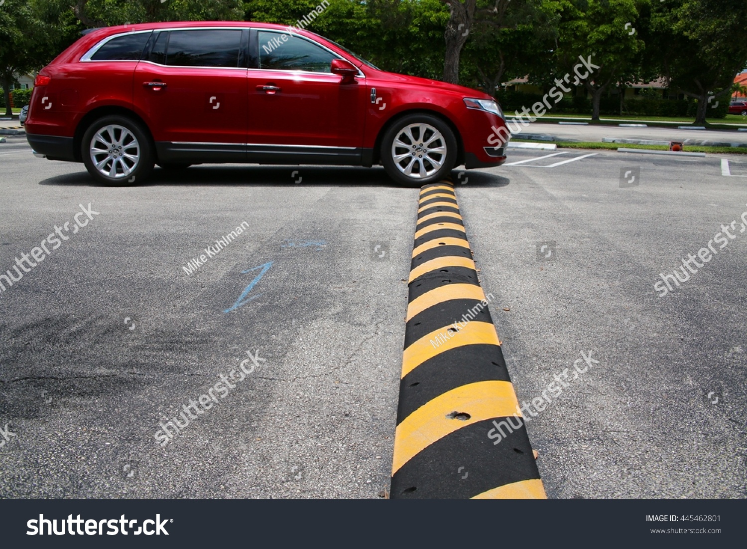 Red Minivan Driving Up to and Just Connecting with Yellow and Black Striped Speed Bump in Parking Lot with Diagonal Striped Spaces and Trees in the Background, Mid-Day #445462801
