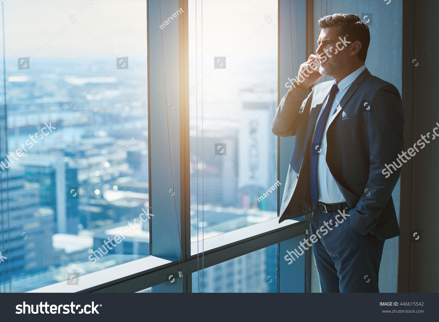 Handsome business CEO with designer stubble, talking confidently on his mobile phone while looking out of large windows in a top floor office at the city below #446615542