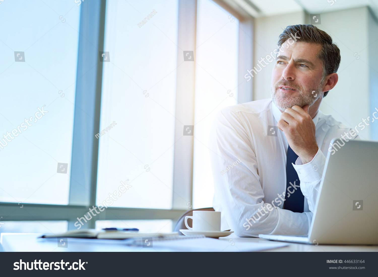 Attractive mature business executive with a stylish short beard, sitting at his office desk and looking out of his window with a thoughtful and optimistic expression #446633164