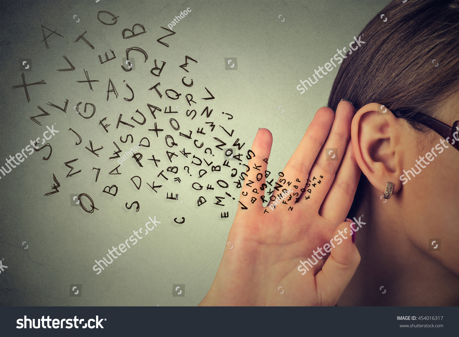 Woman holds her hand near ear and listens carefully alphabet letters flying in isolated on gray wall background  #454016317