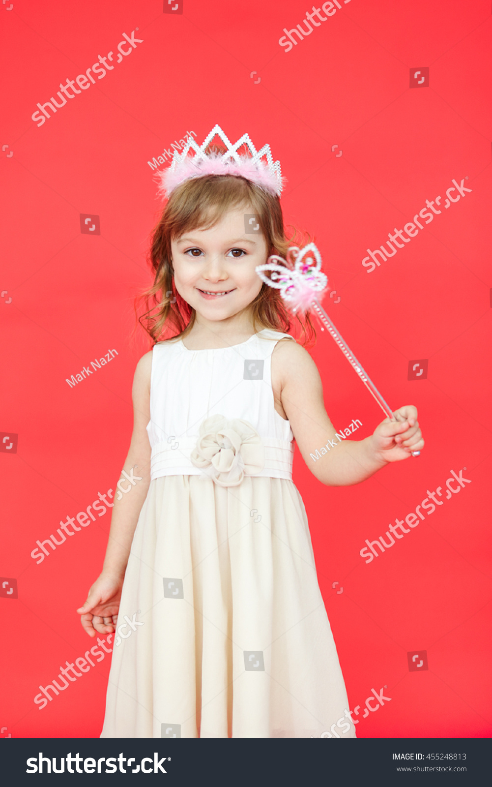 Vertical portrait of beautiful girl dressed in fairy isolated on red background. Cute little kid wearing a crown and white dress holding a magic wand. #455248813