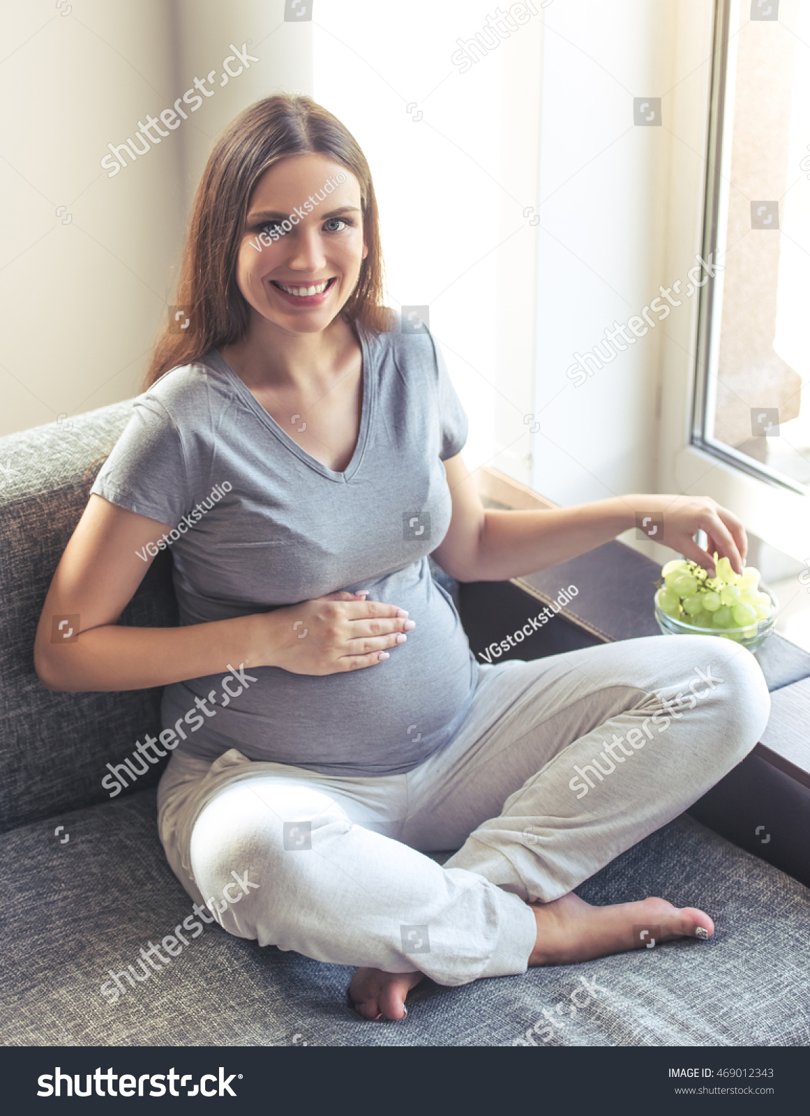 Beautiful pregnant woman is eating grape, holding one hand on her tummy, looking at camera and smiling while sitting on the couch at home #469012343