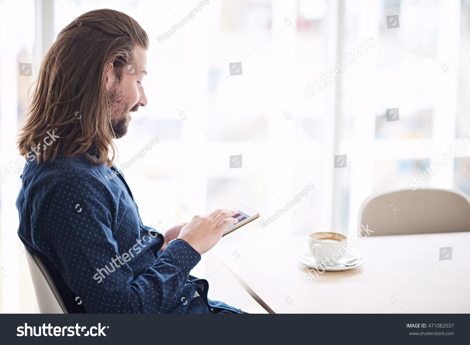 Caucasian man with long brown hair wearing a blue shirt using a tablet while seated at a table in a coffee shop next to large window allowing lots of natural light in. #471082037