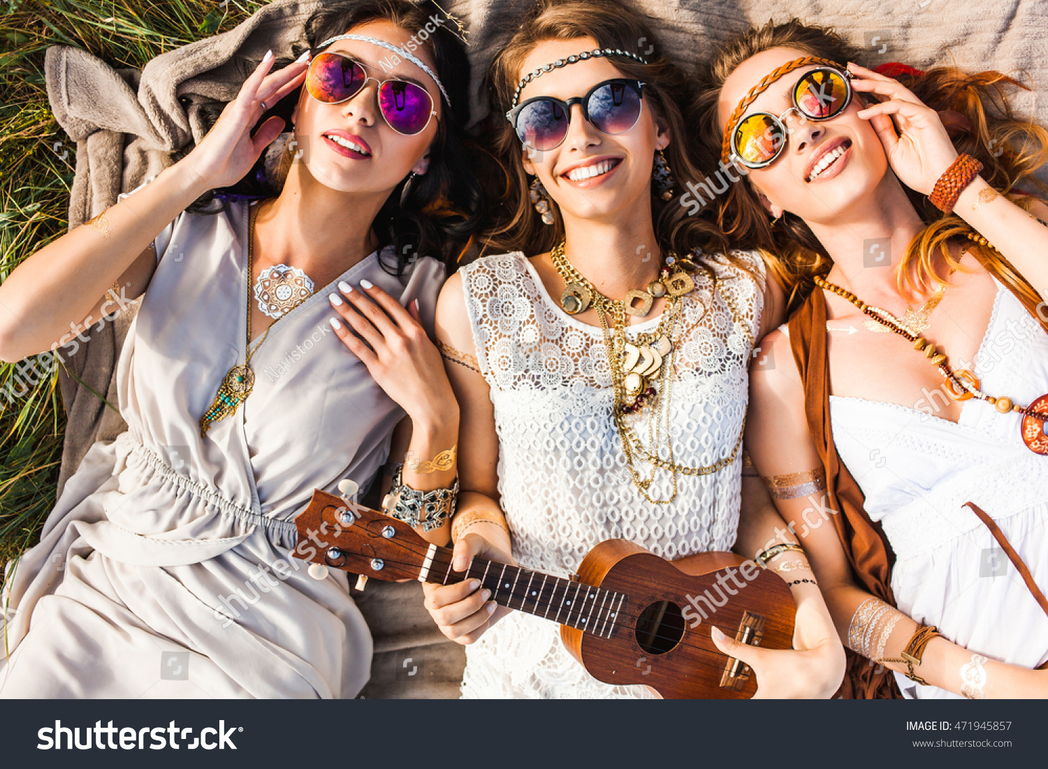 Three cute hippie girl lying on the plaid outdoors, best friends having fun and laughing, play ukulele, sunglasses, feathers in their hair, bracelets, flash tattoo, indie, Bohemia, boho style top view #471945857