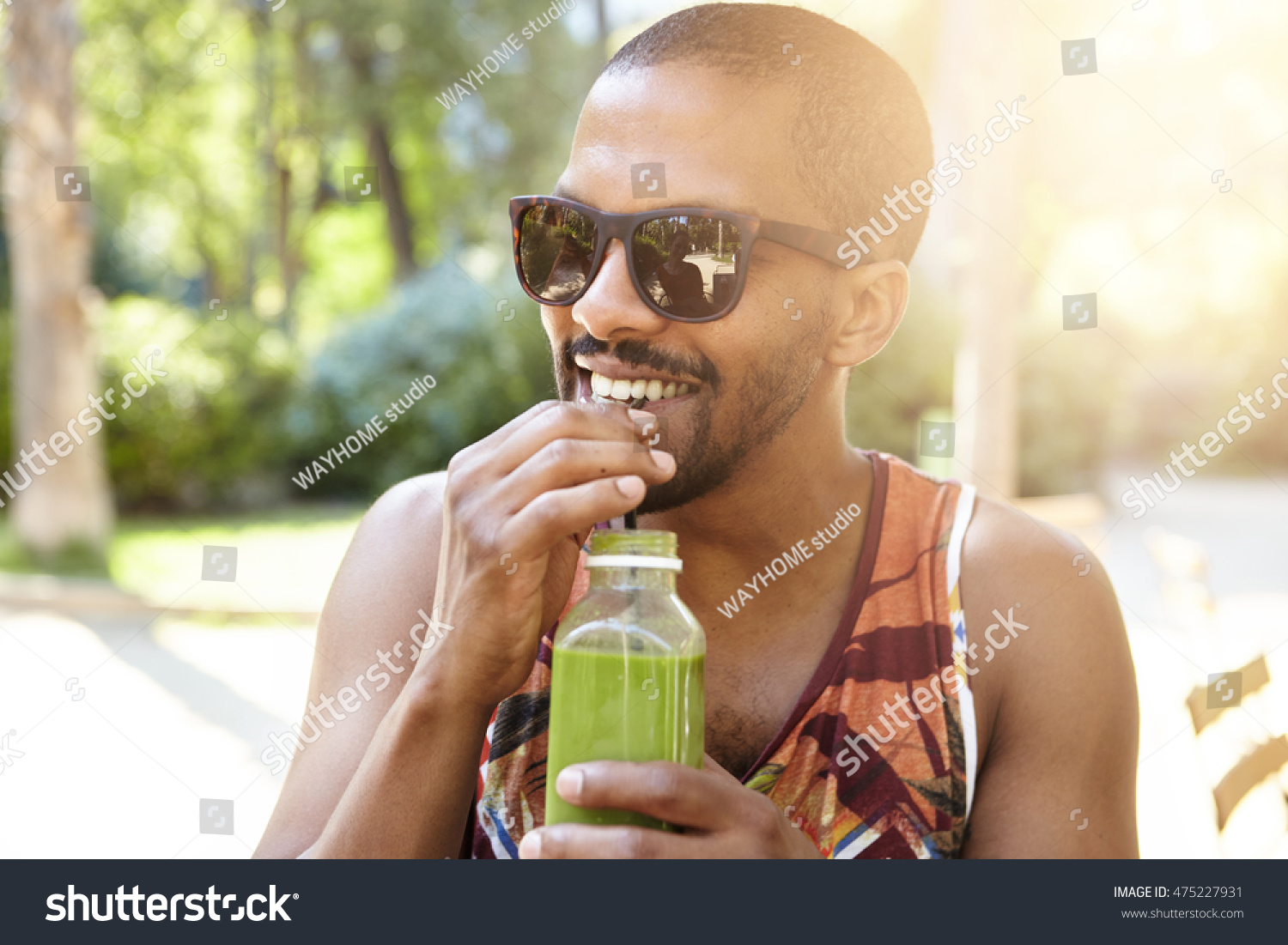 Young attractive African American hipster sipping fresh green smoothie, waiting for his friends at city park on hot summer day, hiding in the shadow of big trees, wearing fashionable sunglasses #475227931