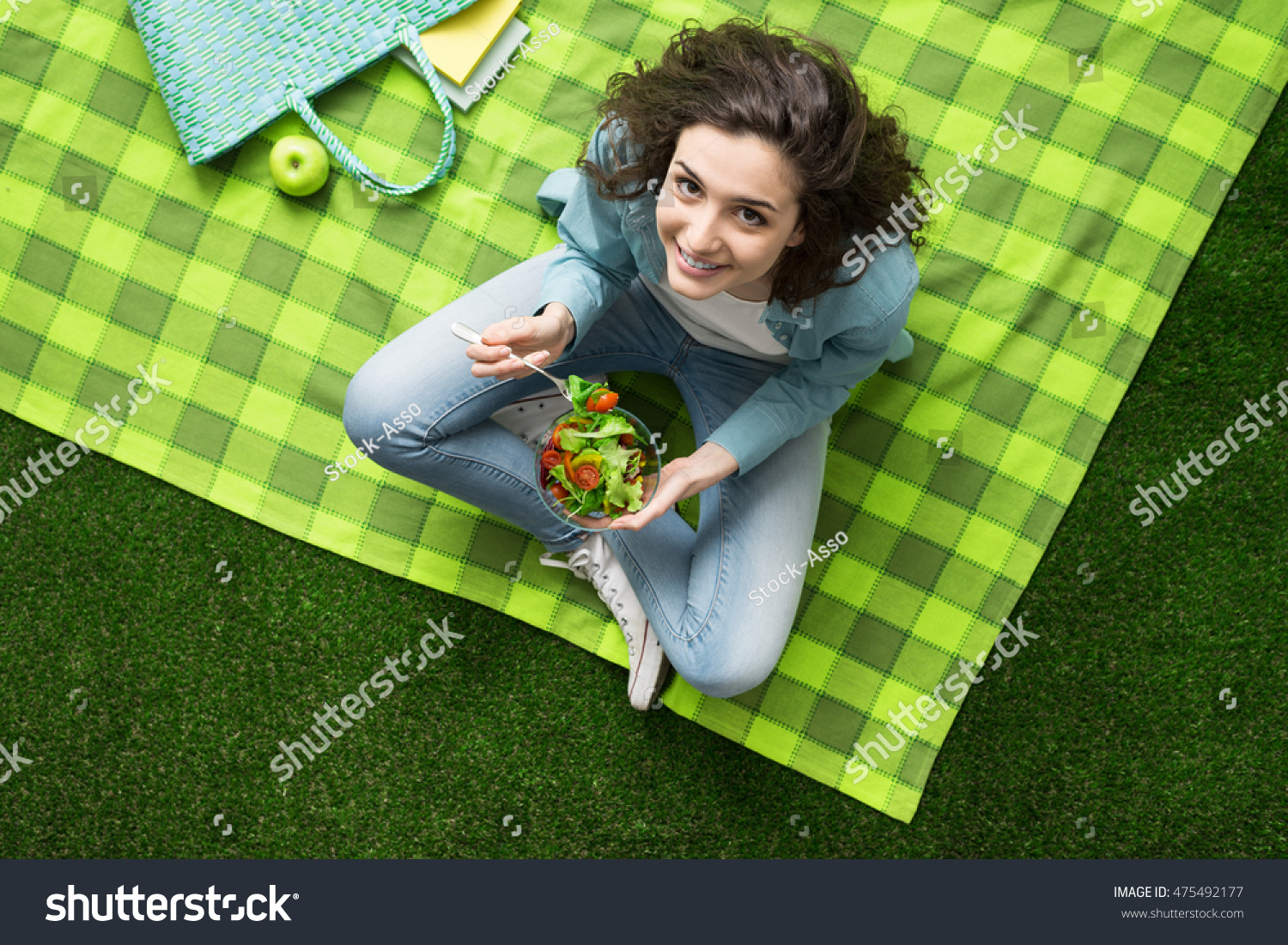 Smiling woman having a relaxing lunch break outdoors, she is sitting on the grass and eating a salad bowl #475492177