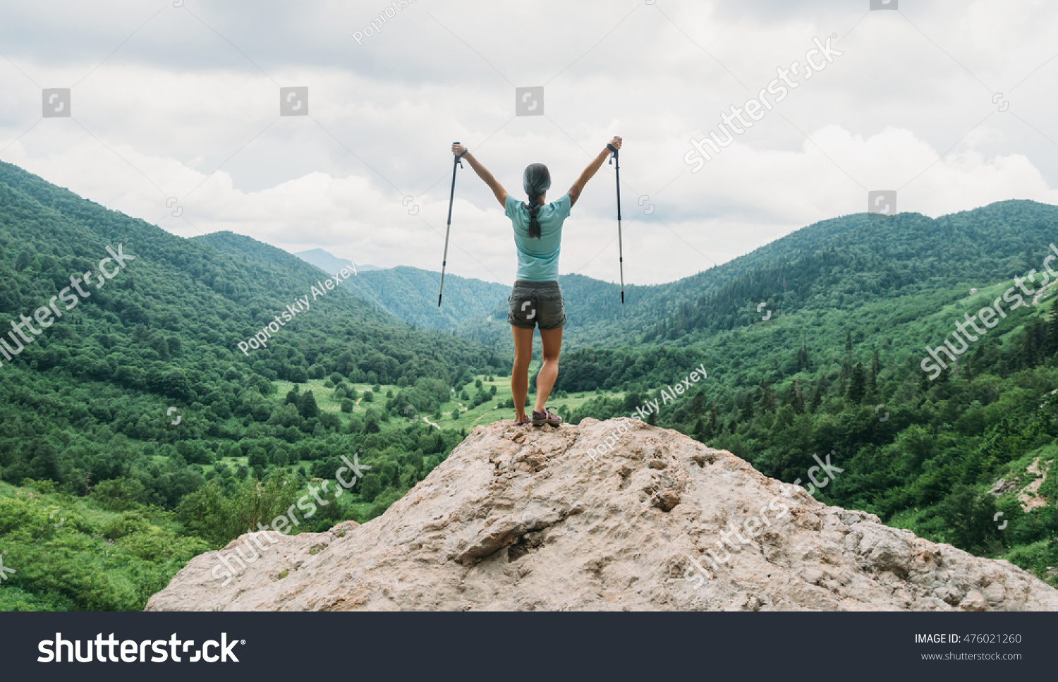 Happy hiker young woman with trekking poles standing on peak of cliff in summer mountains #476021260