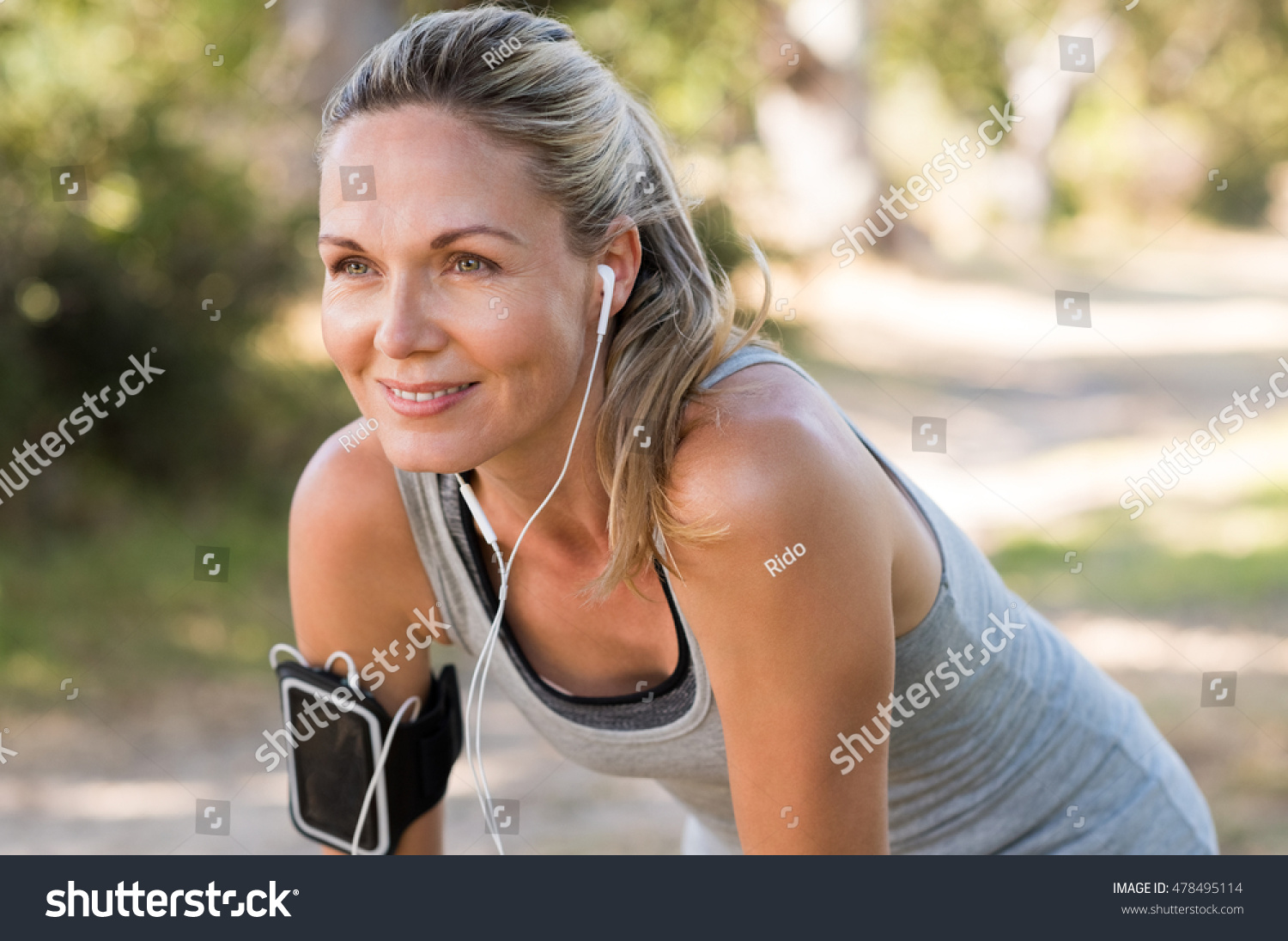 Portrait of athletic mature woman resting after jogging. Beautiful senior blonde woman running at the park on a sunny day. Female runner listening to music while jogging. #478495114