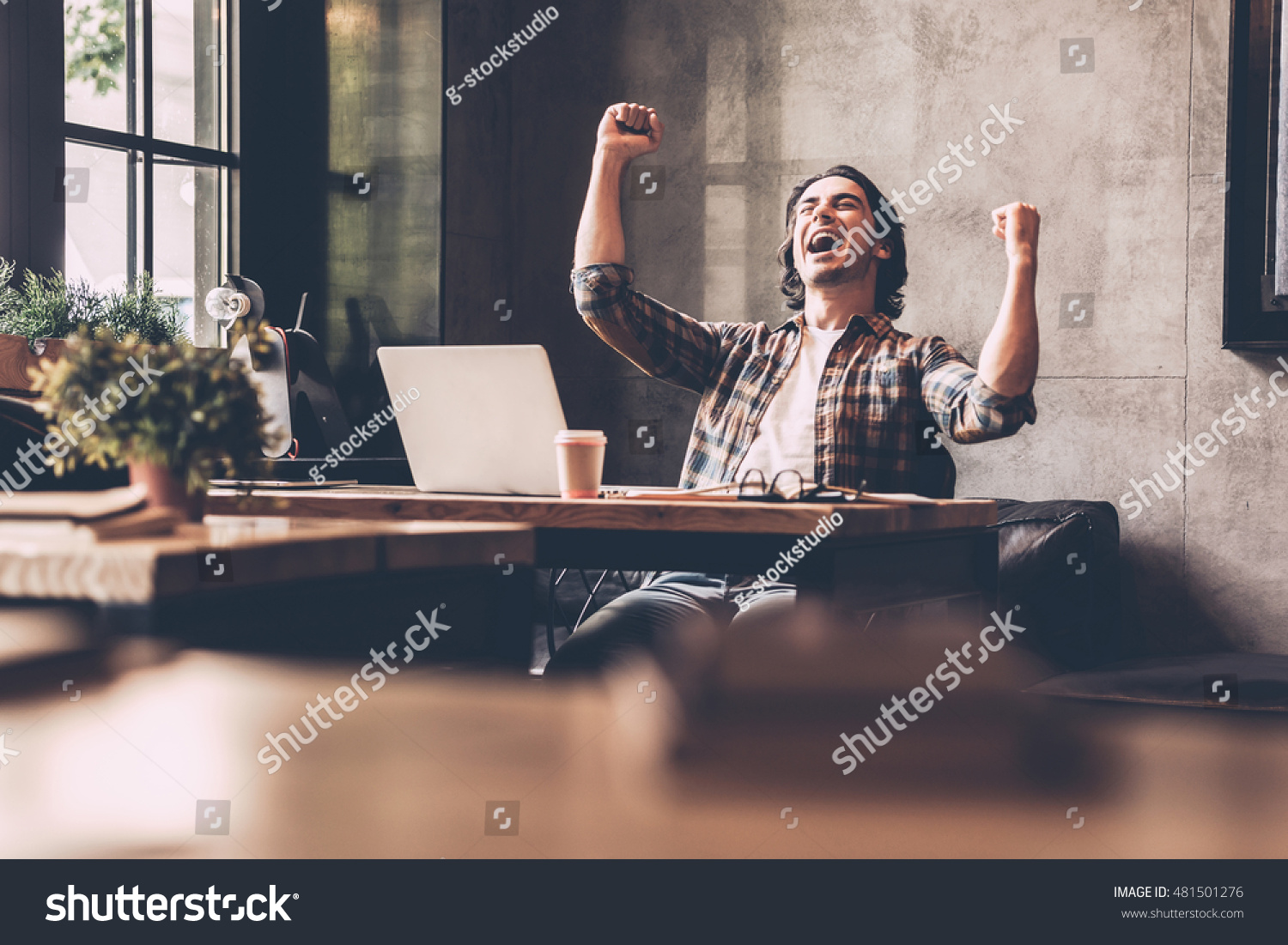 Everyday winner. Cheerful young man in casual wear keeping arms raised and looking happy while sitting at the desk in office  #481501276