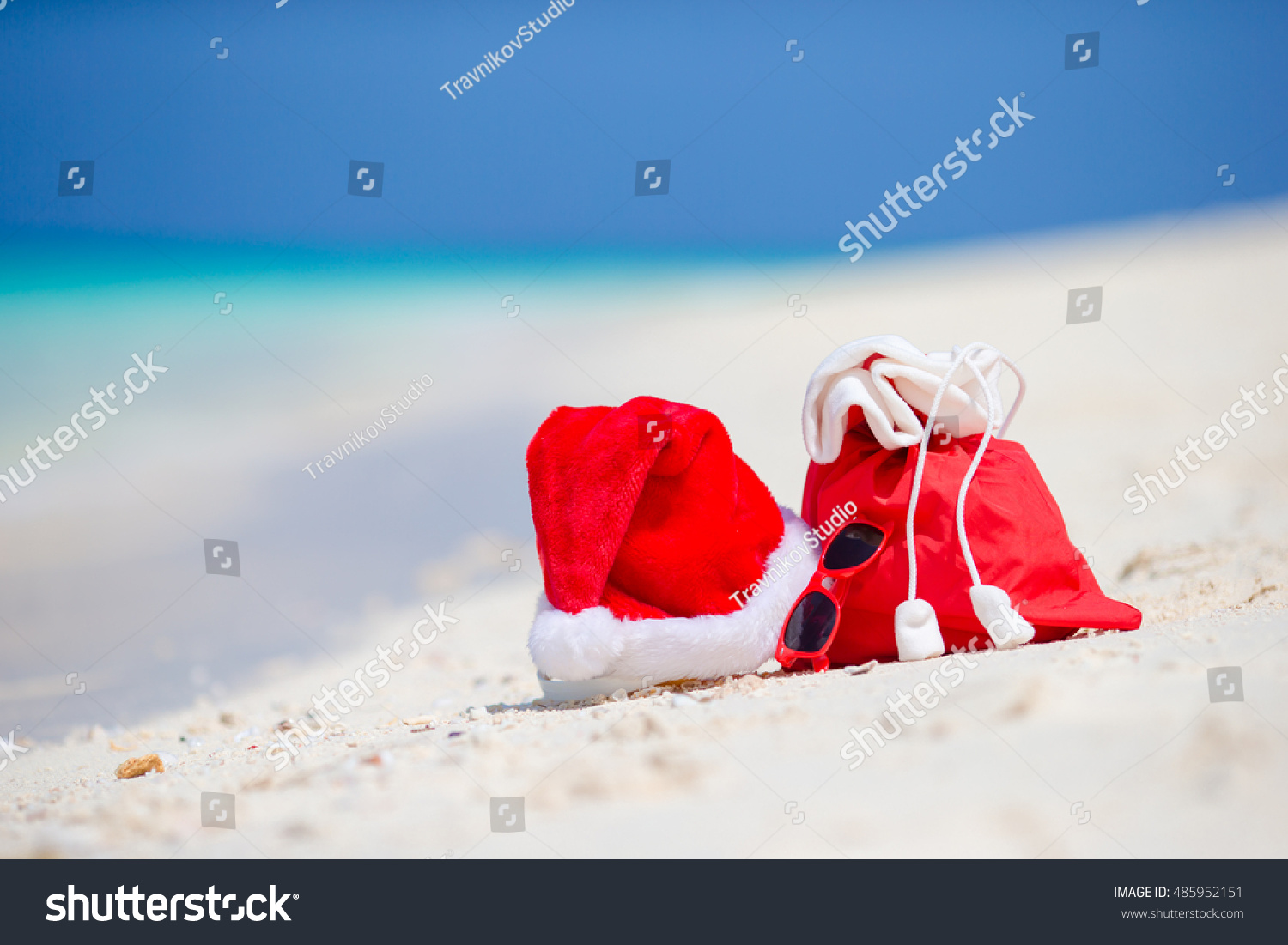 Closeup of red Santa bag and Santa Claus hat on beach. Xmas travel vacation and travel cuprise concept. Beach accessories with Santa Hat on white tropical beach #485952151