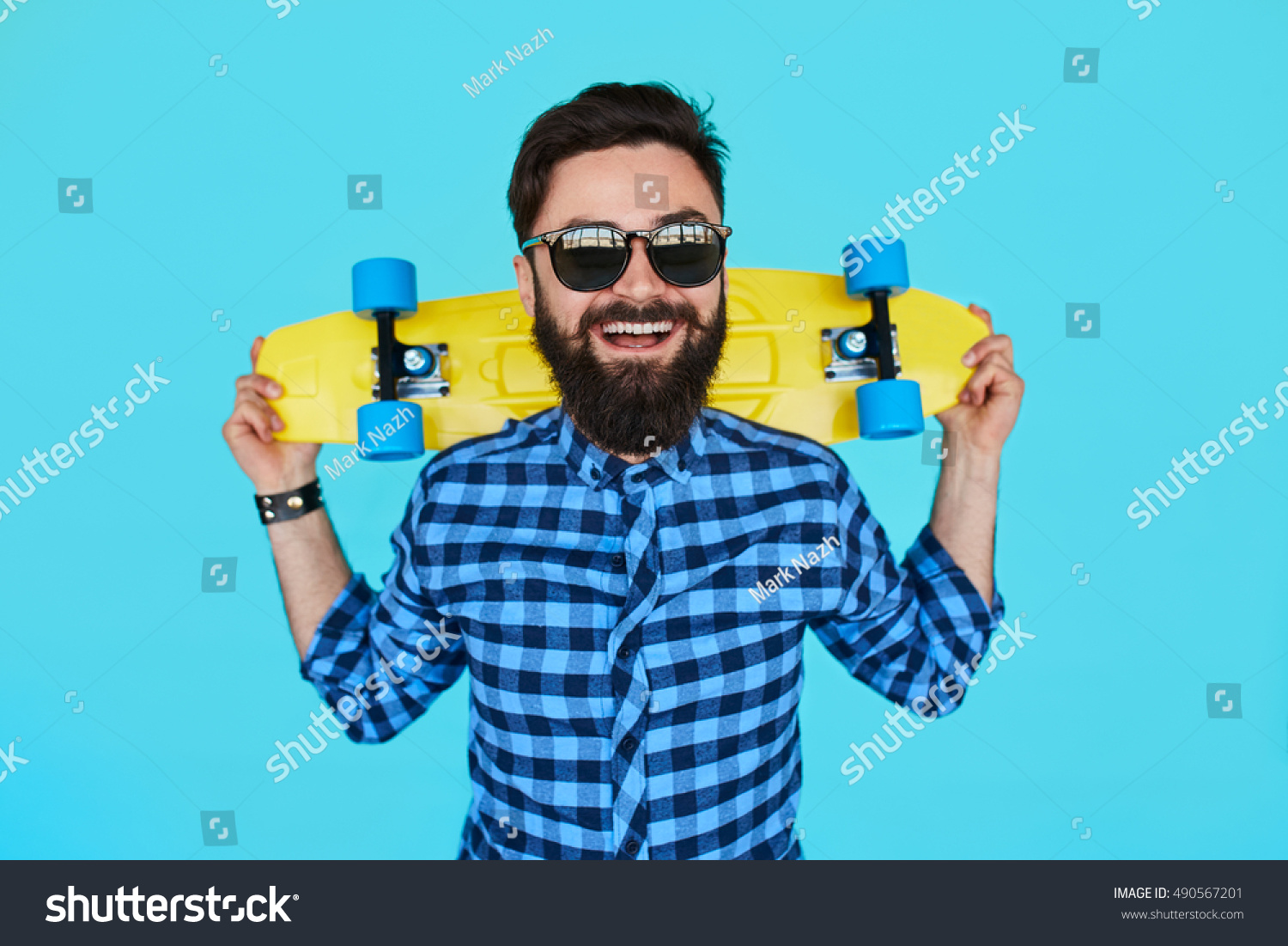 Young hipster bearded man with a yellow skateboard and sunglasses smiling on blue background #490567201