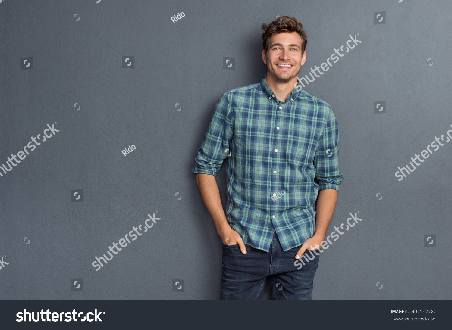 Handsome young man on grey background looking at camera. Portrait of laughing young man with hands in pockets leaning against grey wall. Happy guy smiling. #492562780