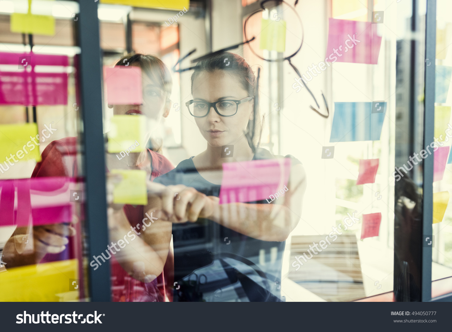 Two business women working together on wall glass with post it stickers. Modern startup office #494050777