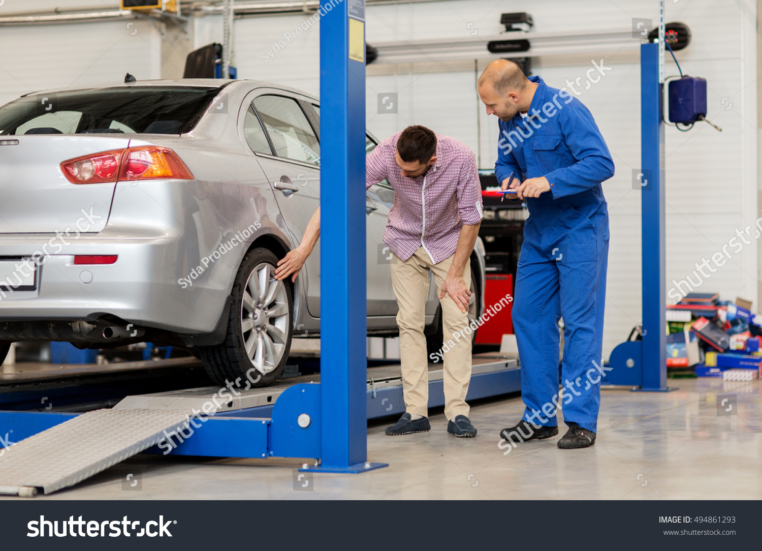 auto service, repair, maintenance and people concept - mechanic with clipboard and man or owner showing wheel at car shop #494861293