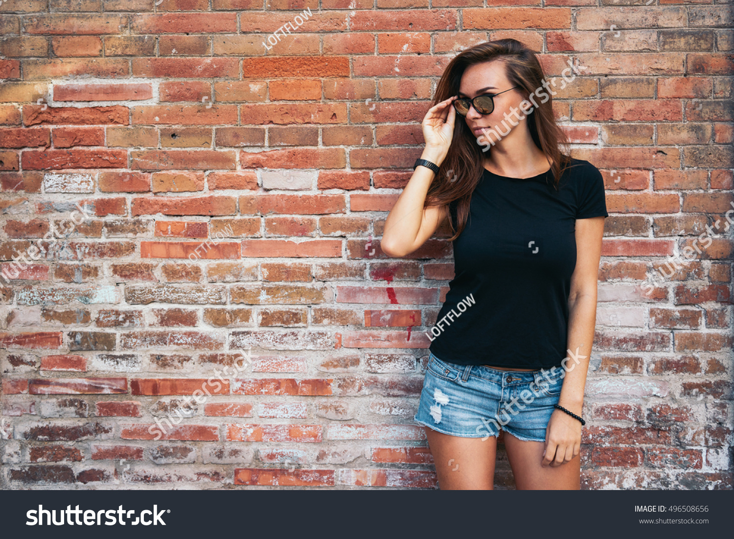 A hipster girl with long brown hair wearing a blank black t-shirt is looking aside while standing on a light blue wooden fence background on a street.Horizontal mock up. Empty space for text o design. #496508656