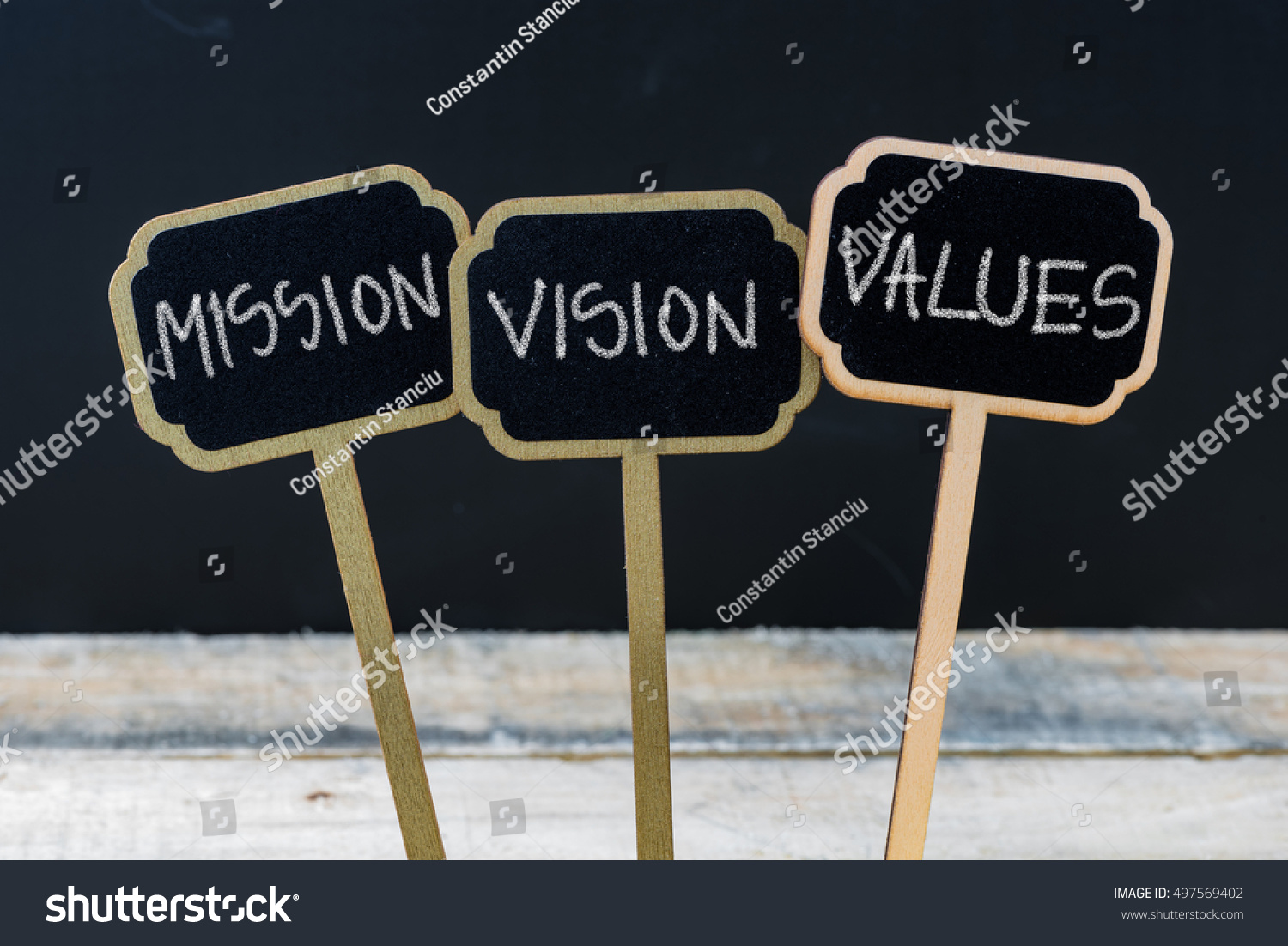 Business message MISSION, VISION, VALUES written with chalk on wooden mini blackboard labels, defocused chalkboard and wood table in background #497569402