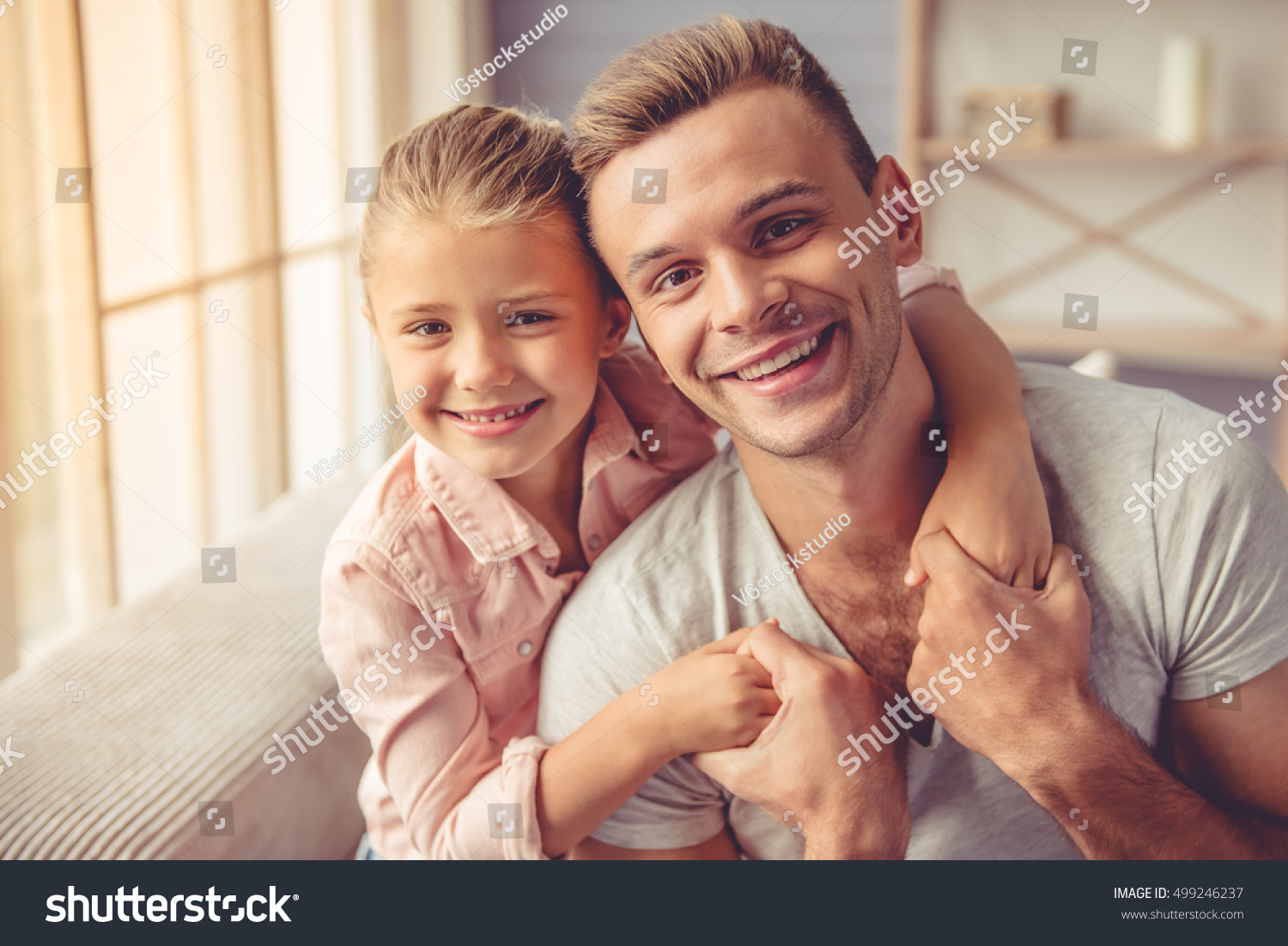 Portrait of handsome young father and his little daughter hugging, looking at camera and smiling while sitting on sofa at home #499246237