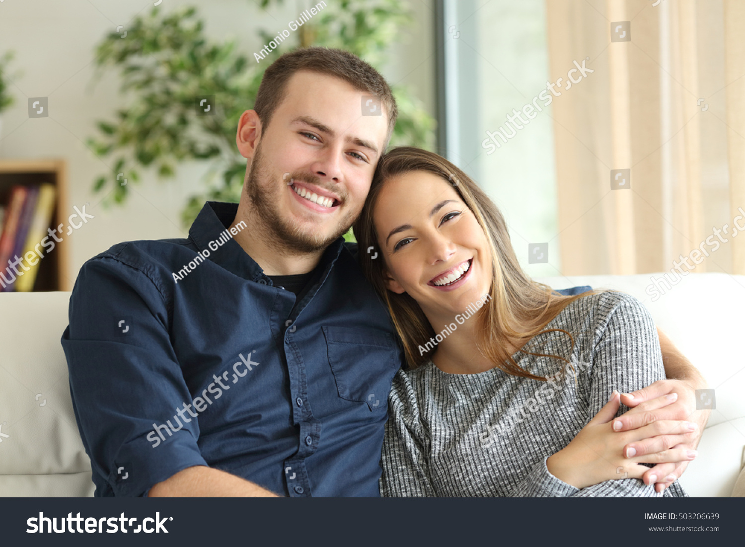 Front view portrait of a happy couple posing and looking at camera sitting on a couch in the living room at home #503206639