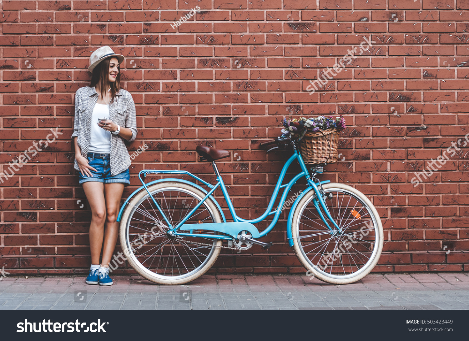Beauty with vintage bike. Beautiful young smiling woman standing near her vintage bicycle with basket full of flowers while she leans against the wall. #503423449