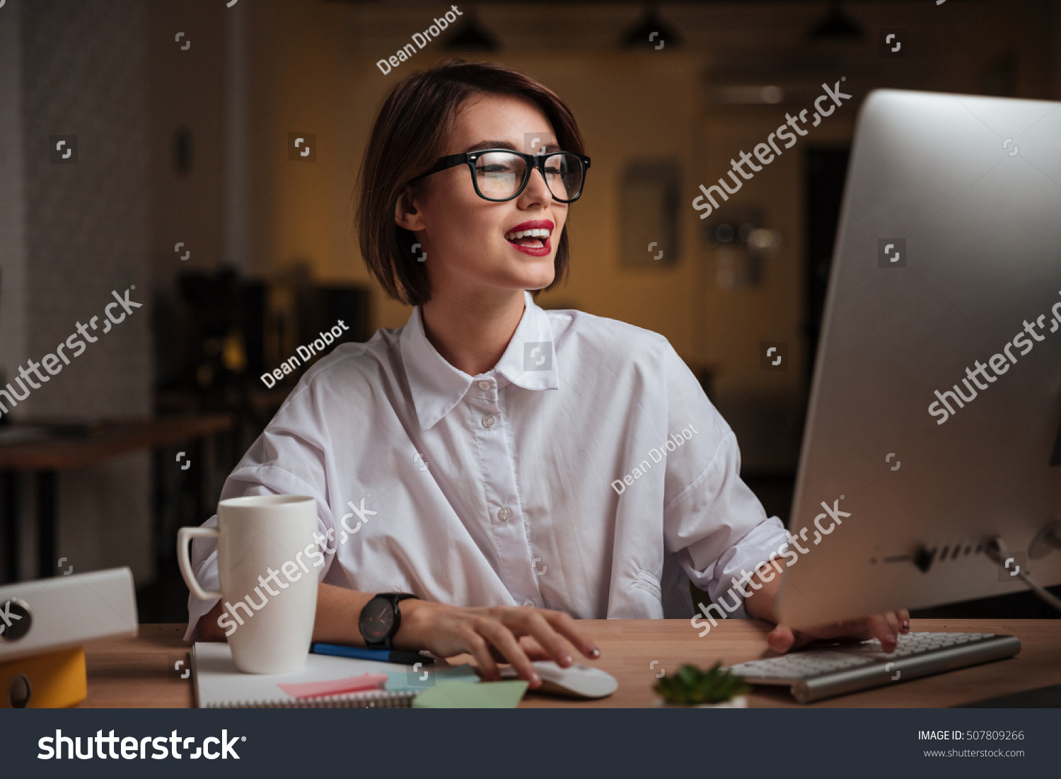 Cheerful young businesswoman in glasses using computer and smiling in office #507809266