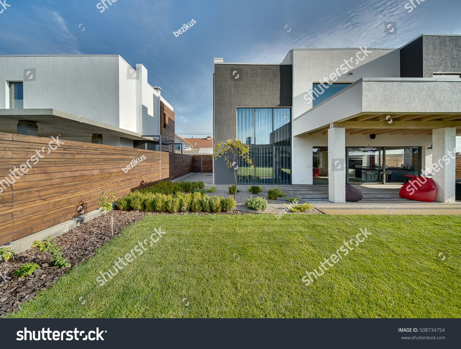 Modern country house with the large lawn and a wooden fence. In front of the house there is a covered terrace with a lounge zone. On the lawn there is a trees and flowerbeds. It is sunny. Outside. #508734754