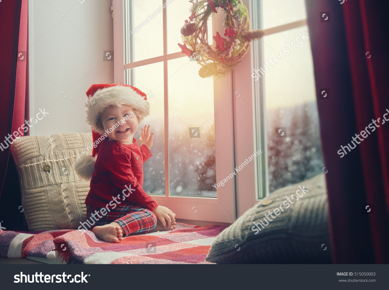 Merry Christmas and happy holidays! Cute little baby girl sitting by the window and looking at the winter forest. Room decorated on Christmas. Kid enjoys the snowfall. #515050003