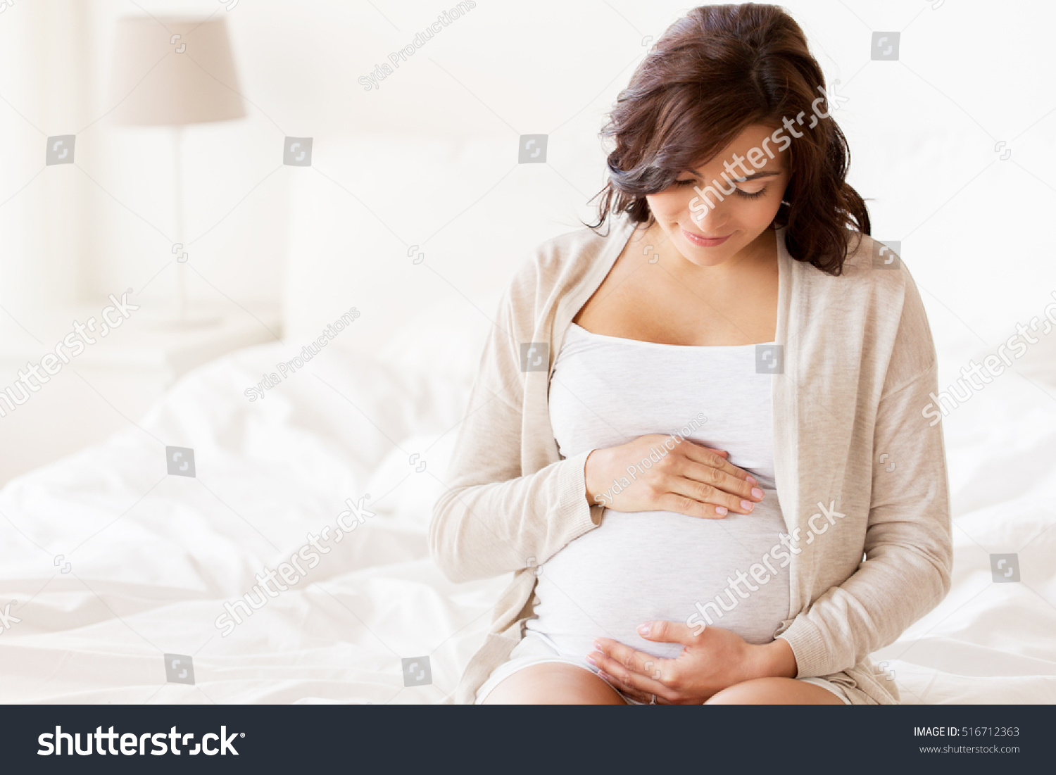 pregnancy, rest, people and expectation concept - happy pregnant woman sitting on bed and touching her belly at home #516712363