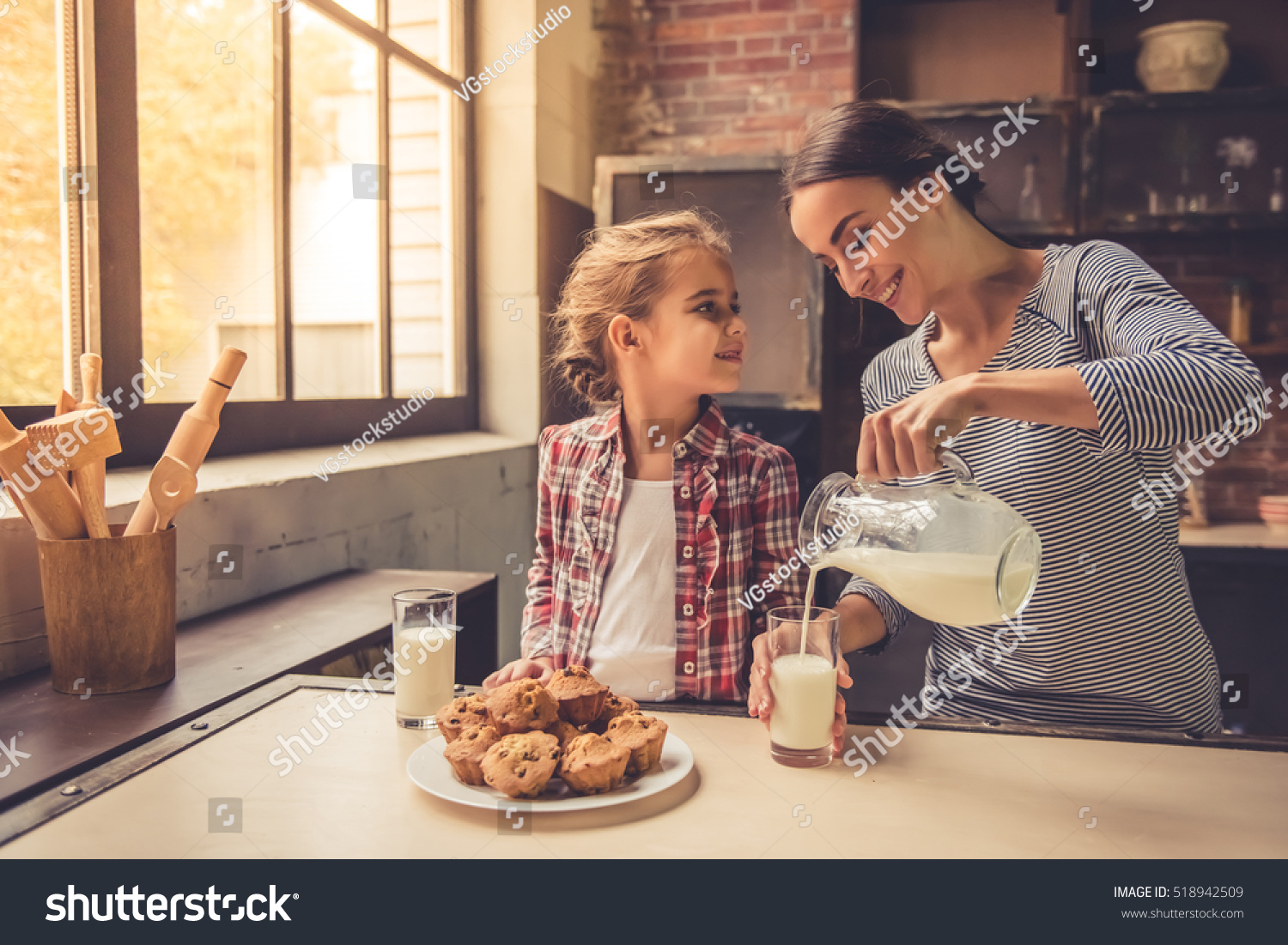 Cute little girl and her beautiful mother are smiling while drinking milk and eating muffins in kitchen. Mother is pouring milk #518942509