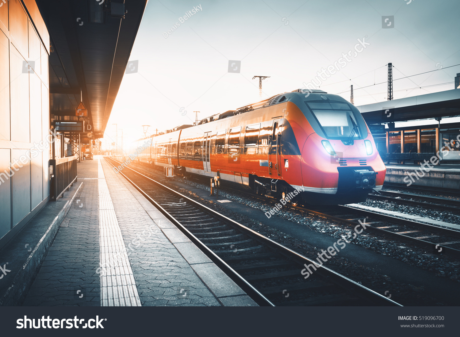 Modern high speed red commuter train at the railway station at sunset. Turning on train headlights. Railroad with vintage toning. Train at railway platform. Industrial landscape. Railway tourism #519096700