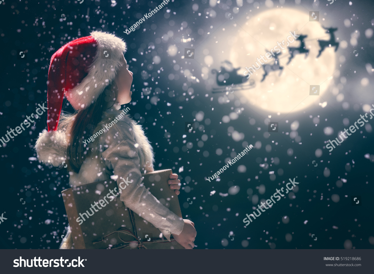 Merry Christmas and happy holidays! Cute little child girl with xmas present. Santa Claus flying in his sleigh against moon sky. Kid enjoy the holiday. Portrait kid with gift on dark background. #519218686