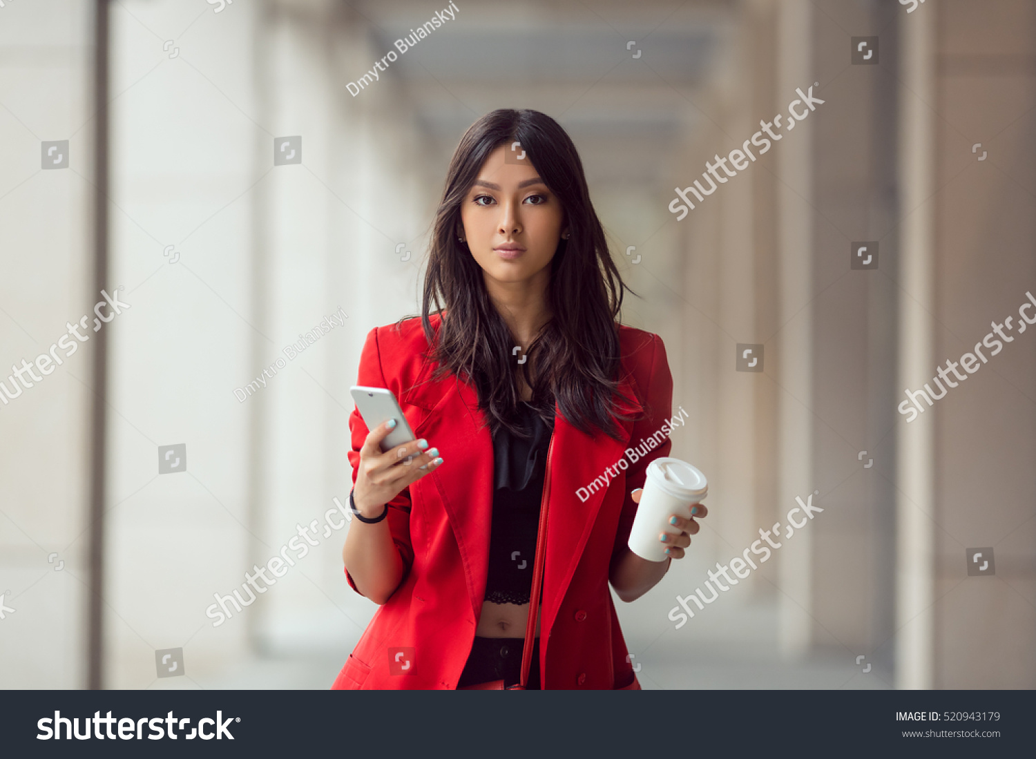 Young Asian woman with smartphone standing against street blurred building background and looking. Fashion business photo of beautiful girl in red casual suite with phone and cup of coffee #520943179