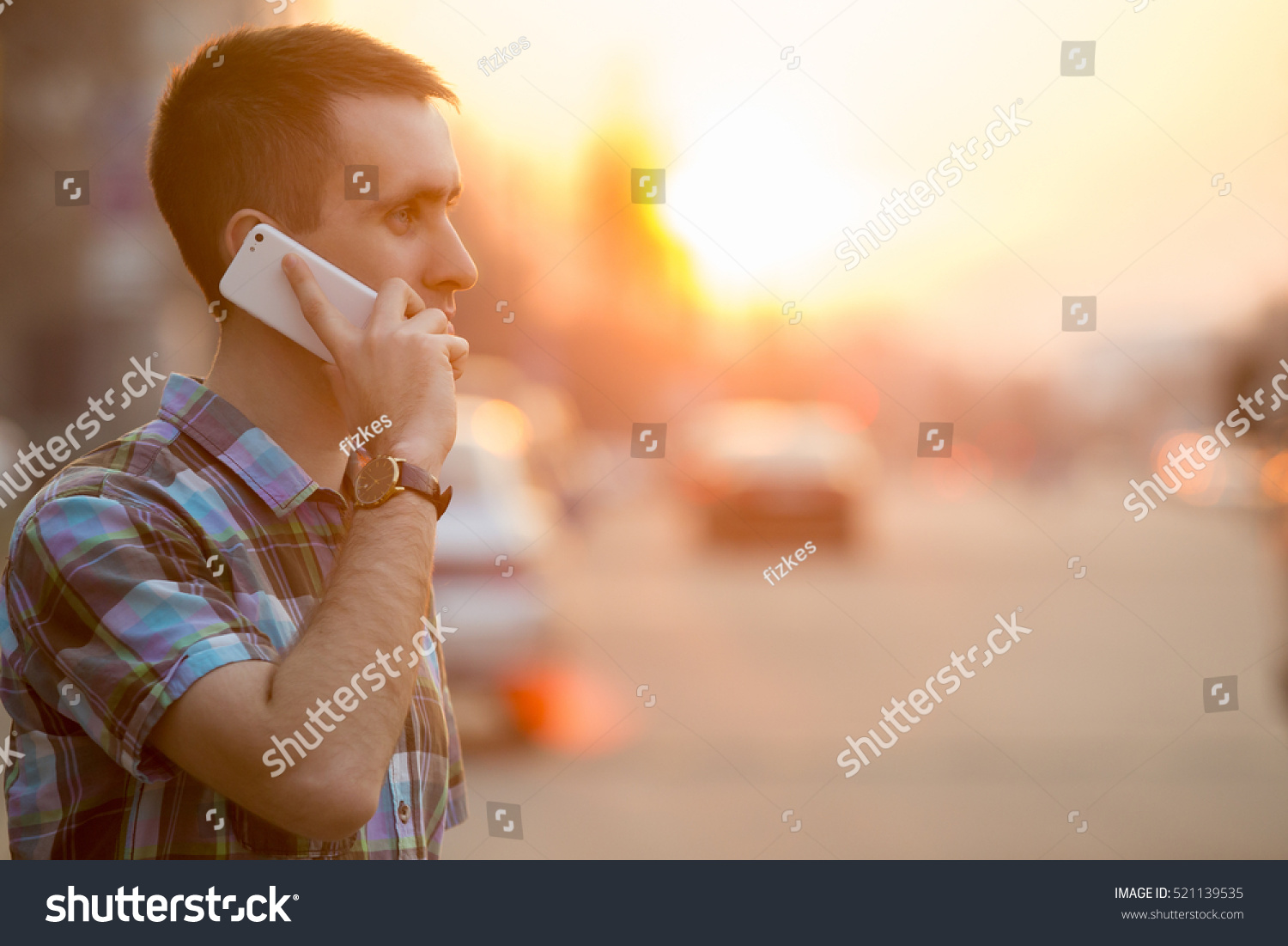 Young man holding mobile phone, using smartphone, making a call, talking on the phone, standing on sunny street with transport traffic on the background #521139535