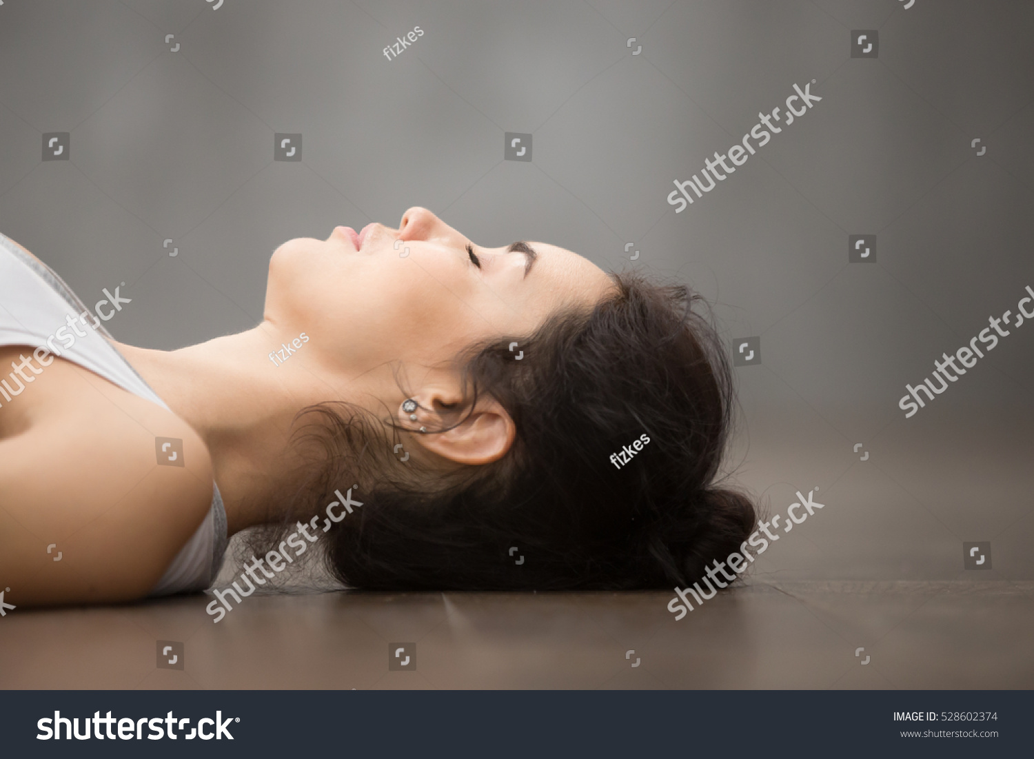 Side view portrait of beautiful young woman working out against grey wall, resting after doing yoga exercises, lying in Shavasana (Corpse or Dead Body Posture), relaxing. Face close up #528602374