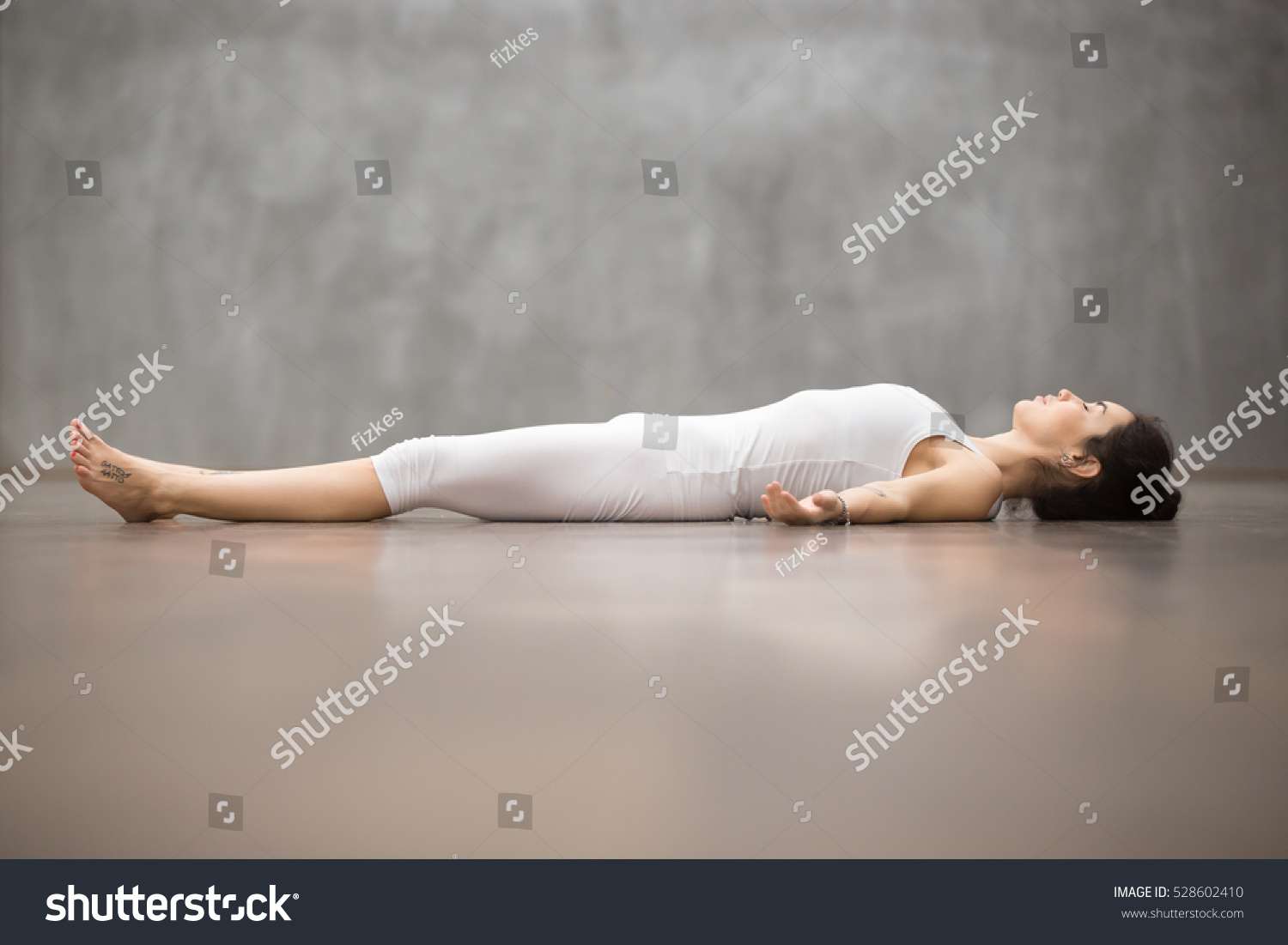Side view portrait of beautiful young woman working out against grey wall, resting after doing yoga exercises, lying in Savasana (Corpse or Dead Body Posture), relaxing. Full length #528602410