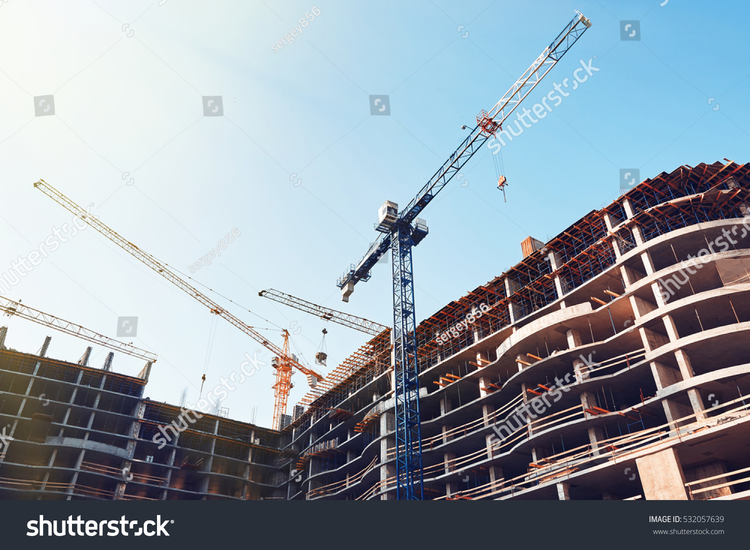 High building under construction. Side with cranes against blue sky with sun glare. #532057639