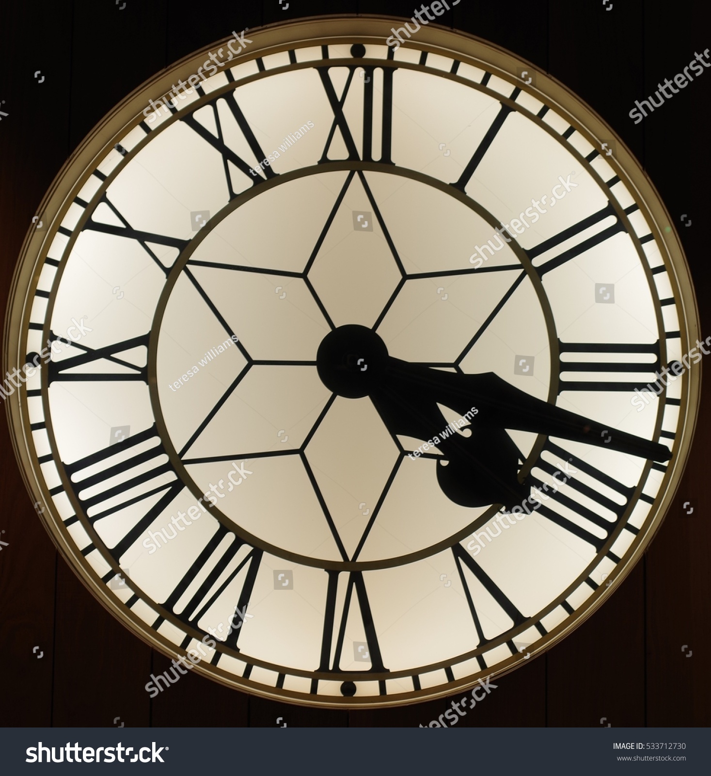 A close up of a circular clock with roman numerals. The time is twenty past four. It has a black background.  The inside of the clock has a star shape. #533712730