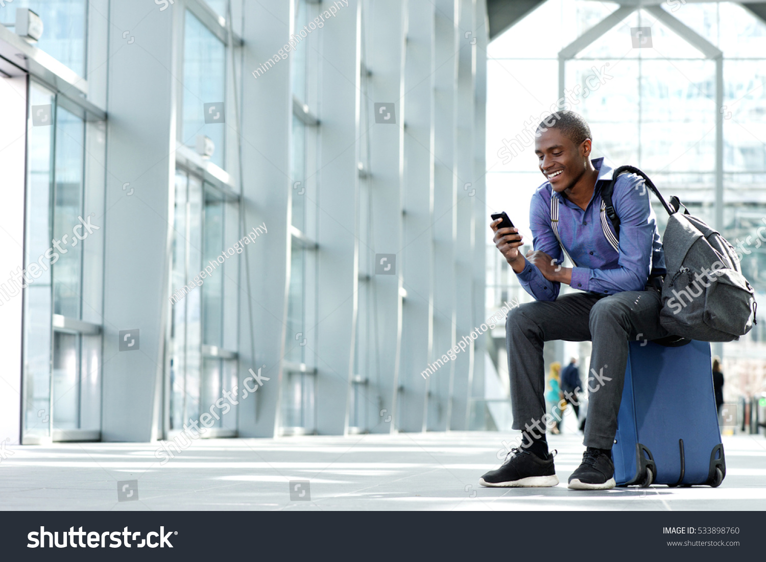 Side portrait of smiling young businessman sitting on suitcase looking at phone #533898760