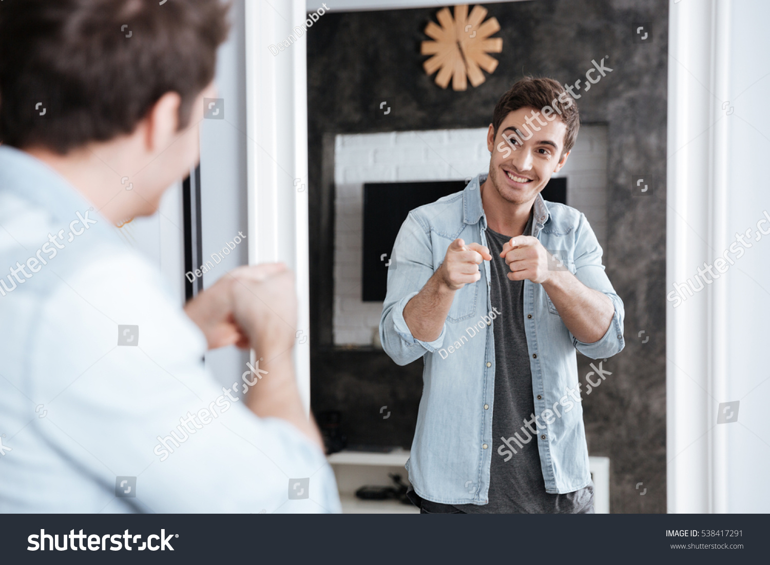 Smiling young man pointing fingers at his mirror reflection while standing at home #538417291
