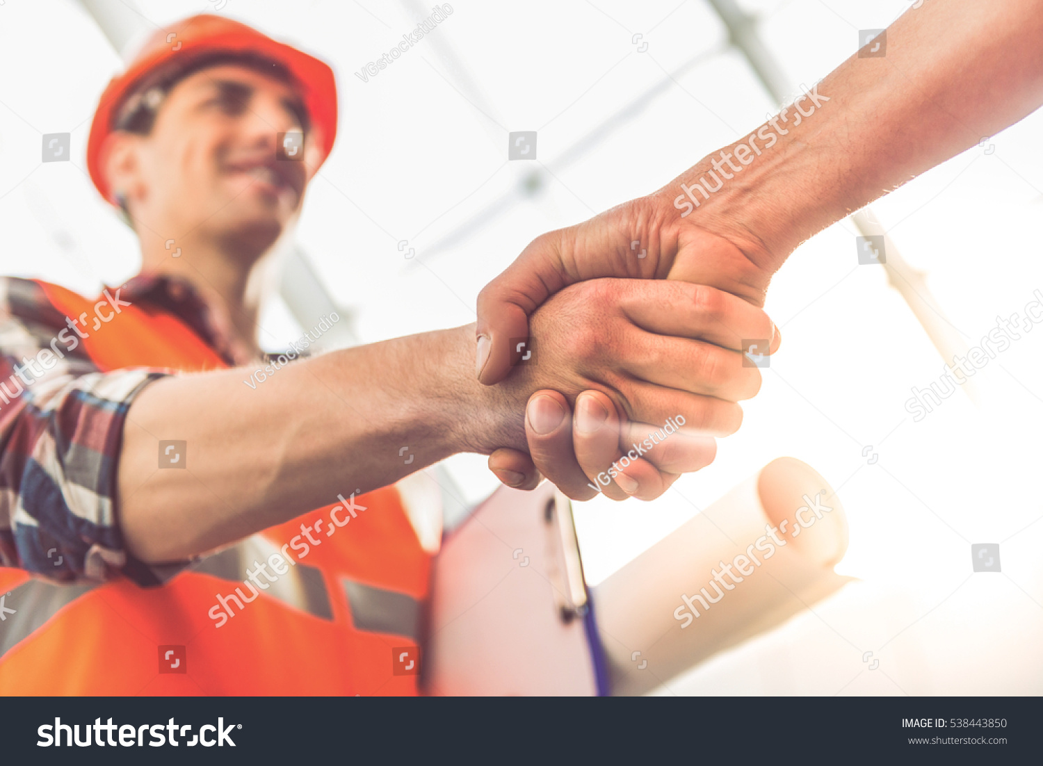 Handsome construction workers in protective helmets and vests are shaking hands while working in the office center #538443850
