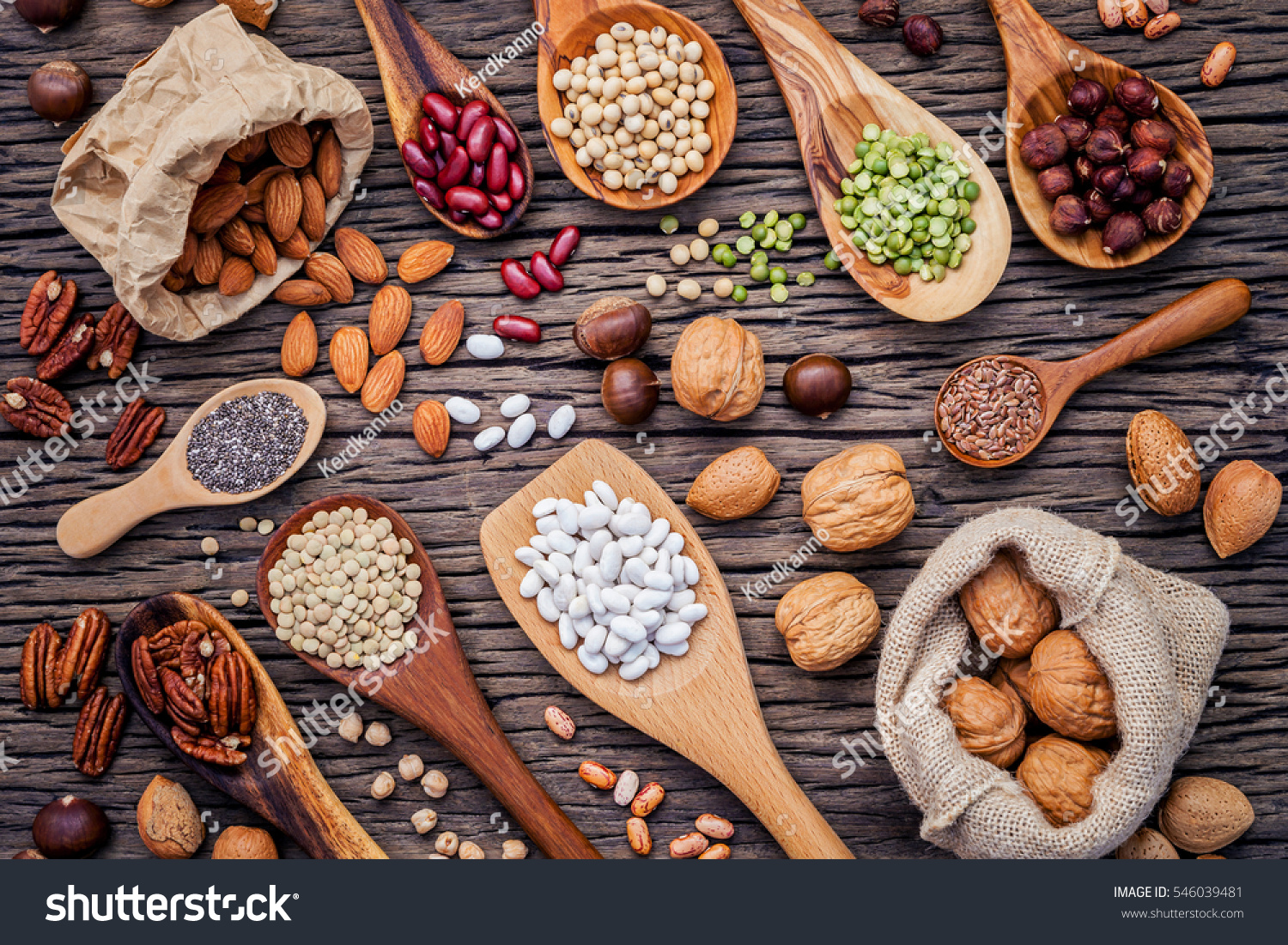 Various legumes and different kinds of nutshells in spoons. Walnuts kernels ,hazelnuts, almond ,brown pinto ,soy beans ,flax seeds ,chia ,chickpea ,red kidney beans and pecan on shabby wooden table. #546039481