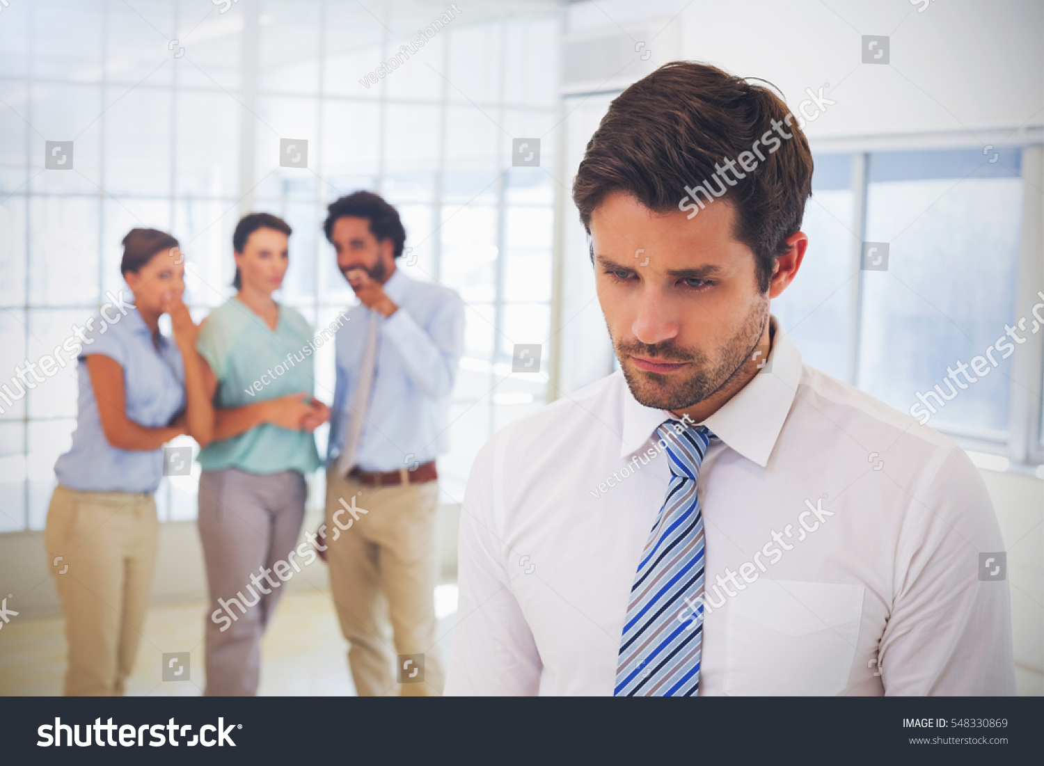 Colleagues gossiping with sad young businessman in foreground at a bright office #548330869