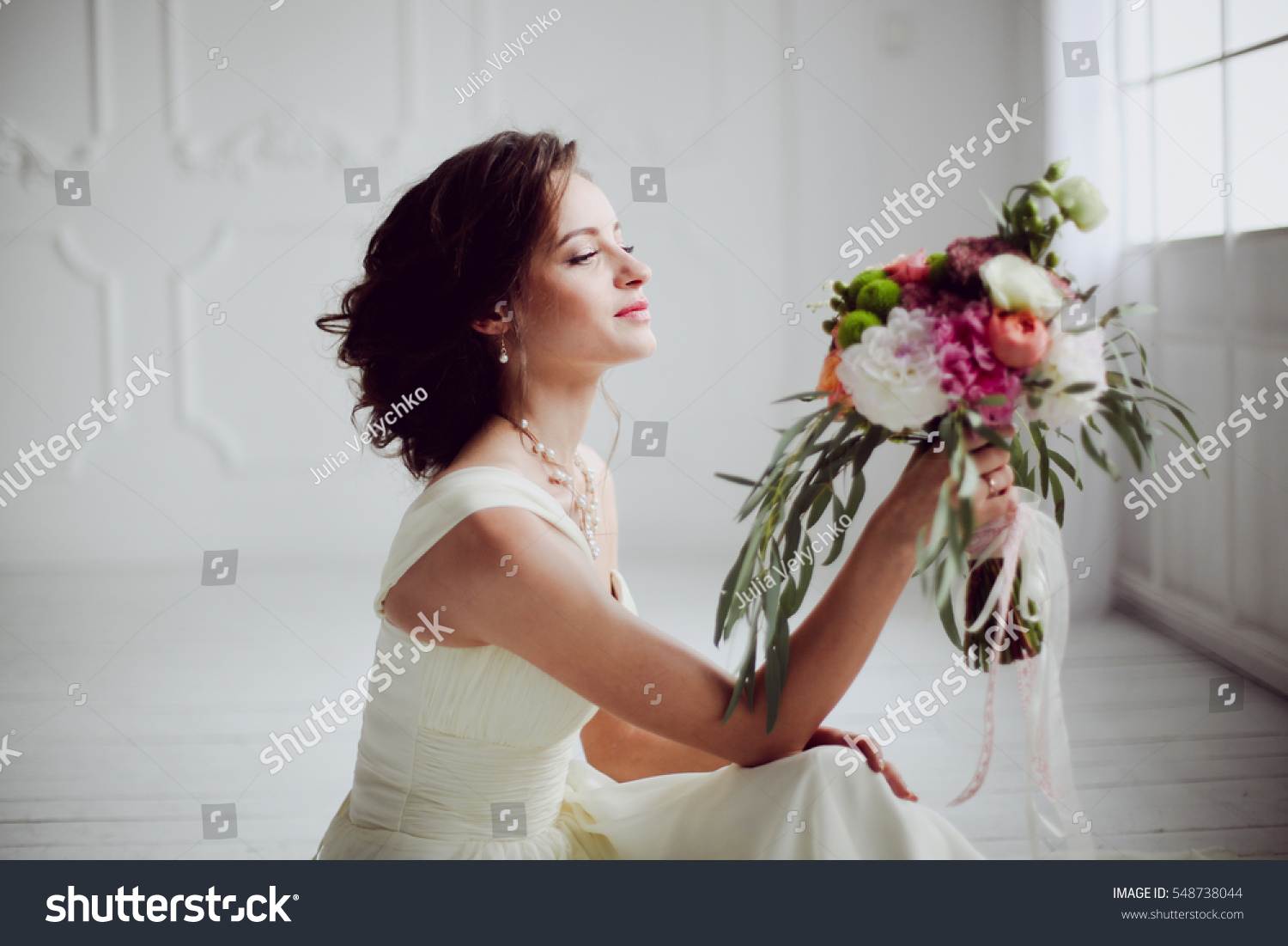 Elegant charming young brunette bride is sitting on white rustic wooden floor in a wedding dress with a big bouquet of flowers, daydreaming,  in the interior Waiting for groom. #548738044