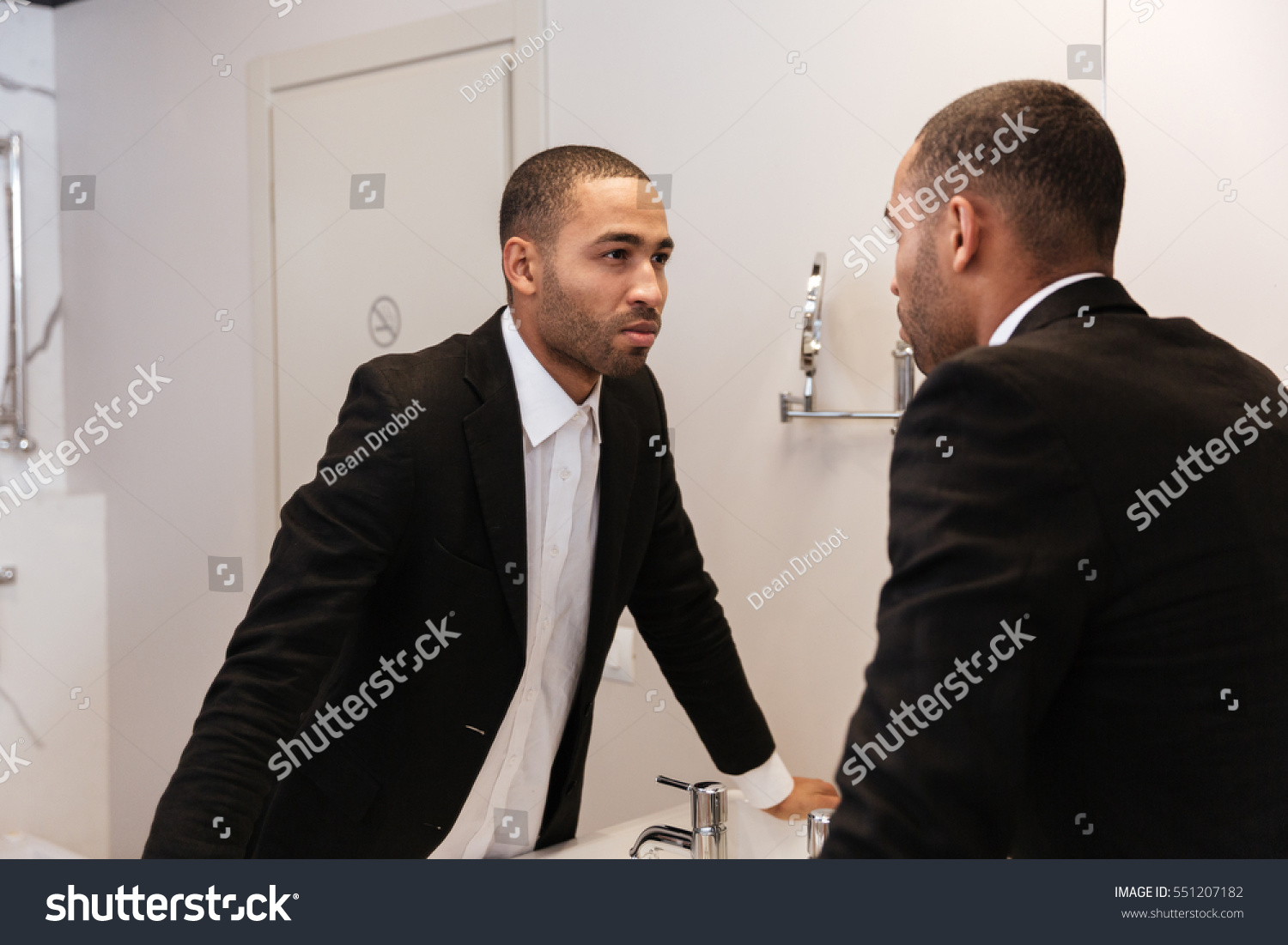Back view of African man in suit looking at mirror in bathroom in hotel room #551207182